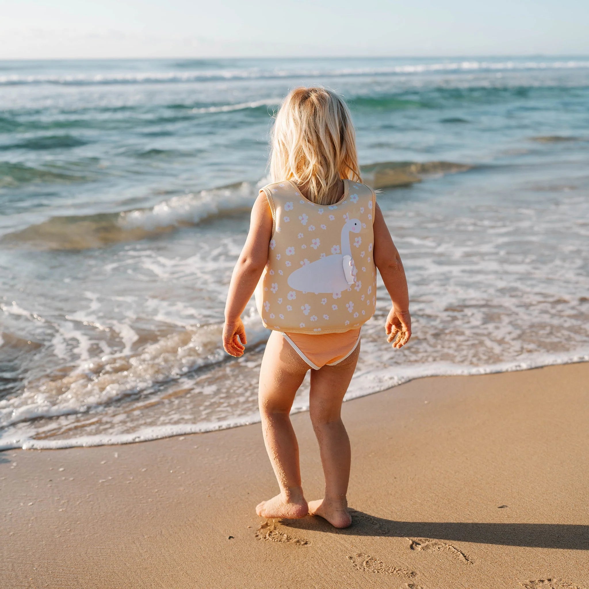 A young child with blonde hair stands at the edge of the beach, wearing a SUNNYLIFE Kids Swim Vest Princess Swan/Buttercup and beige swim bottoms, looking out at the ocean waves. The vest features an adjustable safety strap and is made from durable neoprene. The sky is clear and the sun is shining.