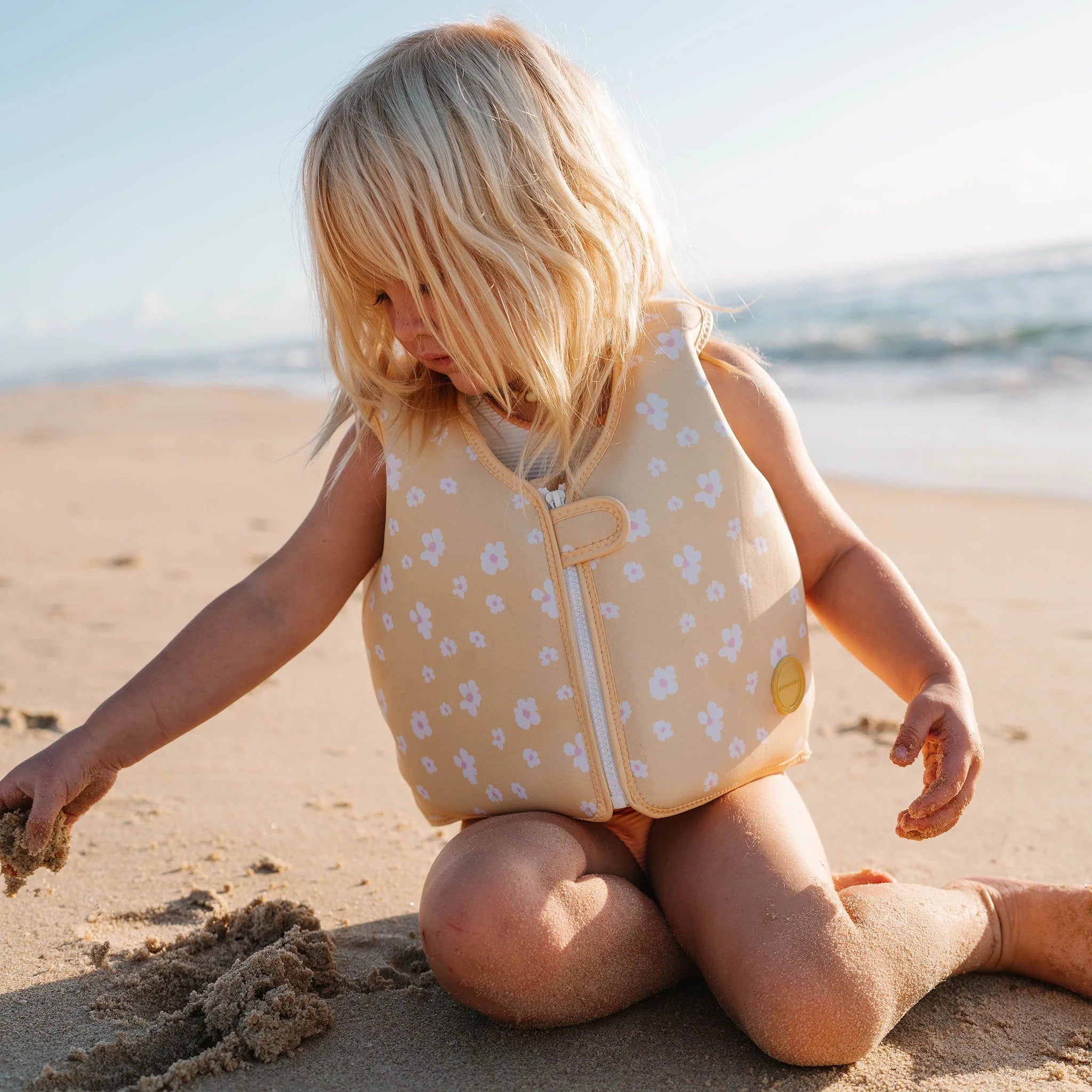 A young child with blonde hair wears a SUNNYLIFE Kids Swim Vest Princess Swan/Buttercup, made of durable neoprene and adorned with white flowers, while sitting on a sandy beach. The child is playing with the sand, and the ocean is visible in the background under a clear sky. An adjustable safety strap ensures their swimming abilities are safe.