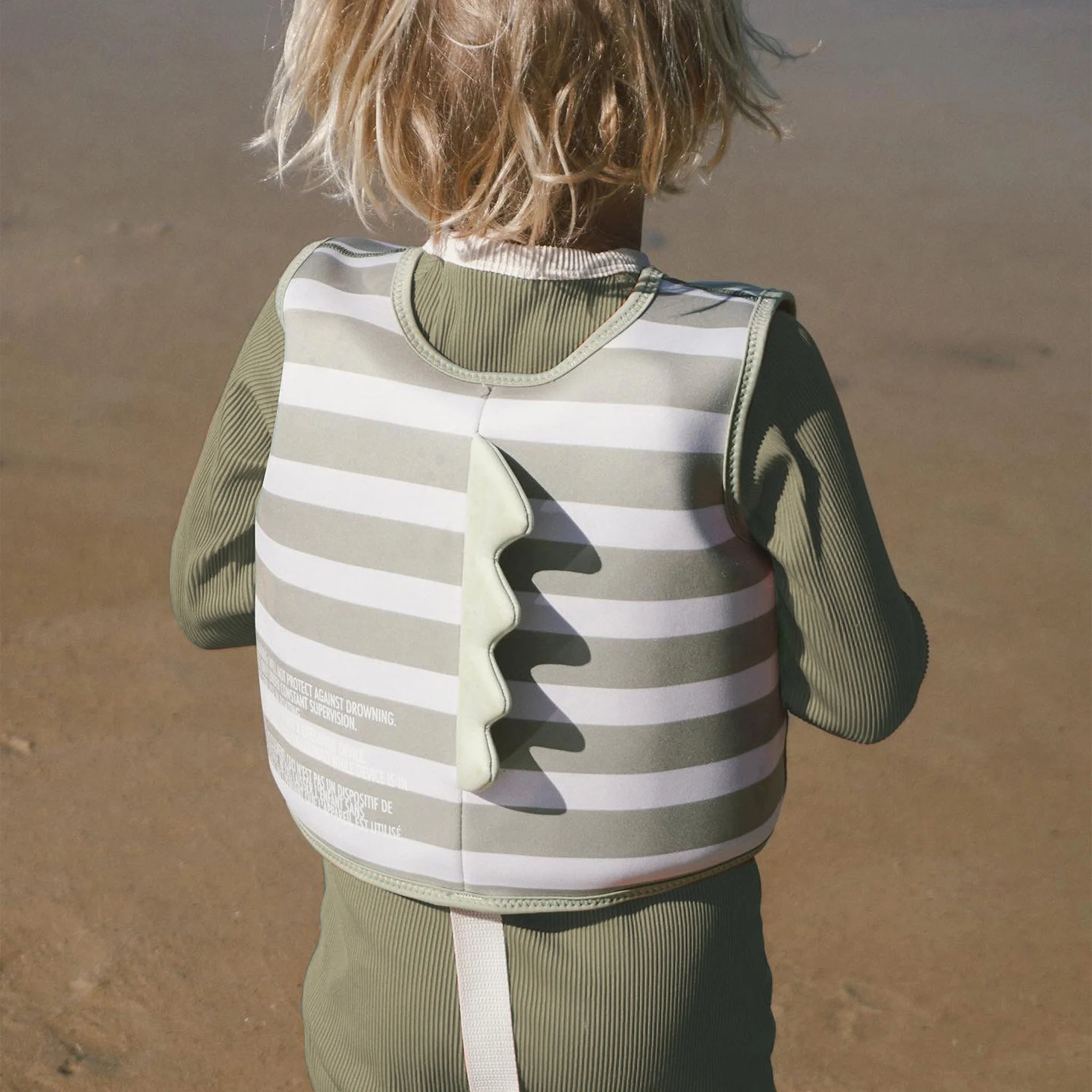 A young child with blonde hair, wearing a green long-sleeve outfit and a striped SUNNYLIFE Kids Swim Vest in Into the Wild/Khaki with a fin-like attachment on the back, stands facing away from the camera on a sandy beach with water in the background. The swim vest features an adjustable safety strap, perfect for developing swimming abilities.