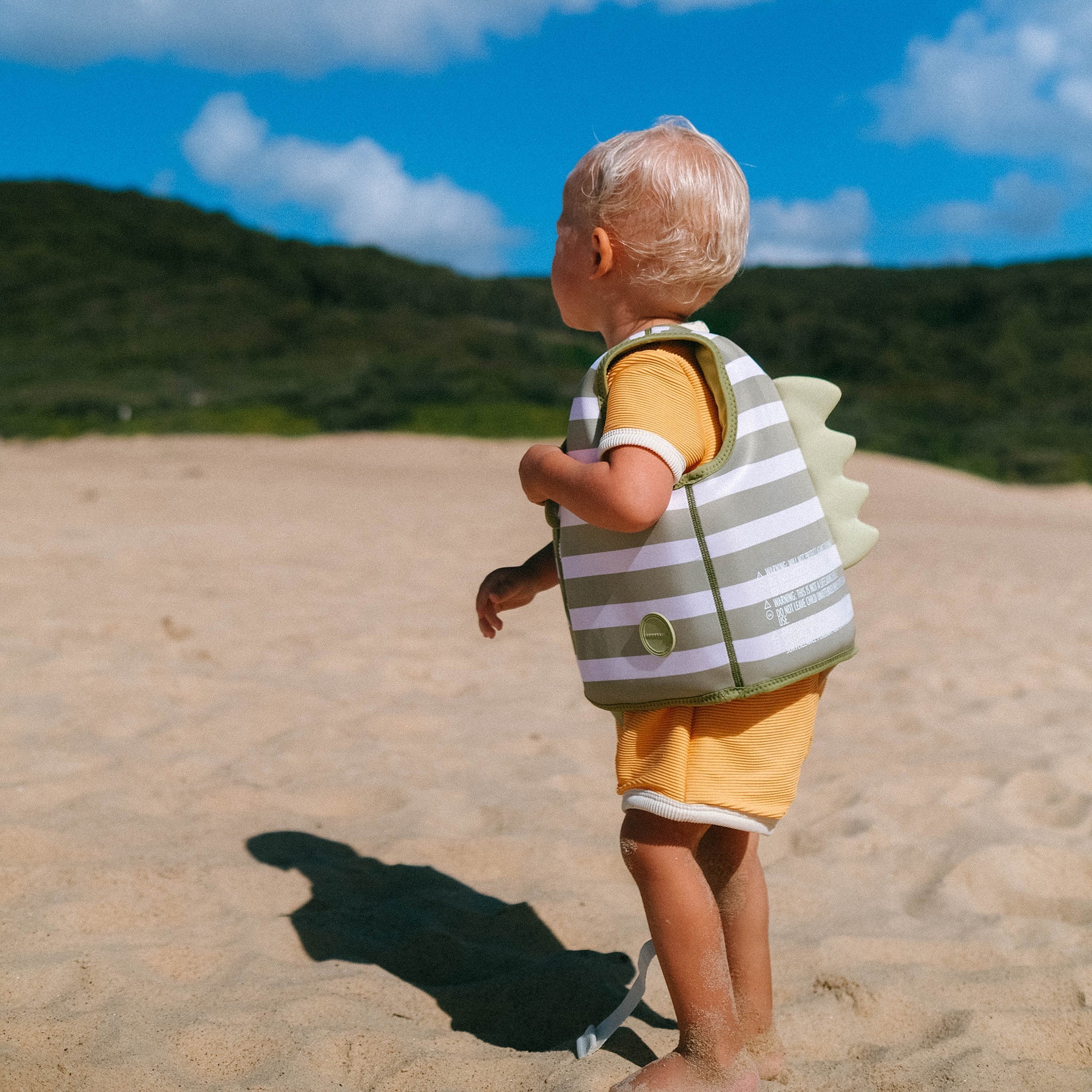 A toddler with blonde hair stands on a sandy beach, wearing a SUNNYLIFE Kids Swim Vest Into the Wild/Khaki and a yellow outfit. The child, ready to develop swimming abilities, faces away, looking toward grassy hills in the background under a partly cloudy sky. The swim vest features an adjustable safety strap for comfort.