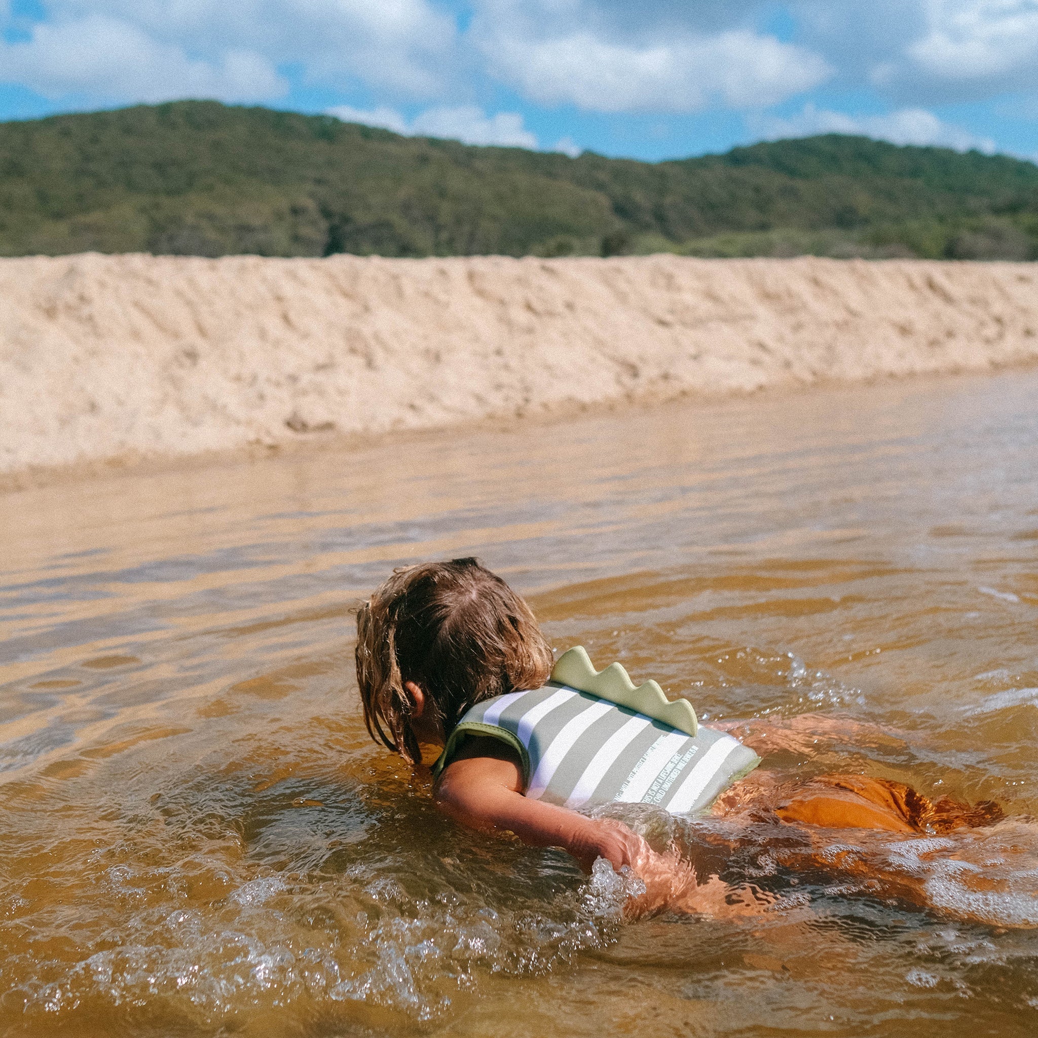 A young child wearing a SUNNYLIFE Kids Swim Vest in the Into the Wild/Khaki design with an adjustable safety strap swims in shallow water near a sandy shore. The child's hair is wet and they are facing away from the camera. In the background, there are green hills under a blue sky with scattered clouds.