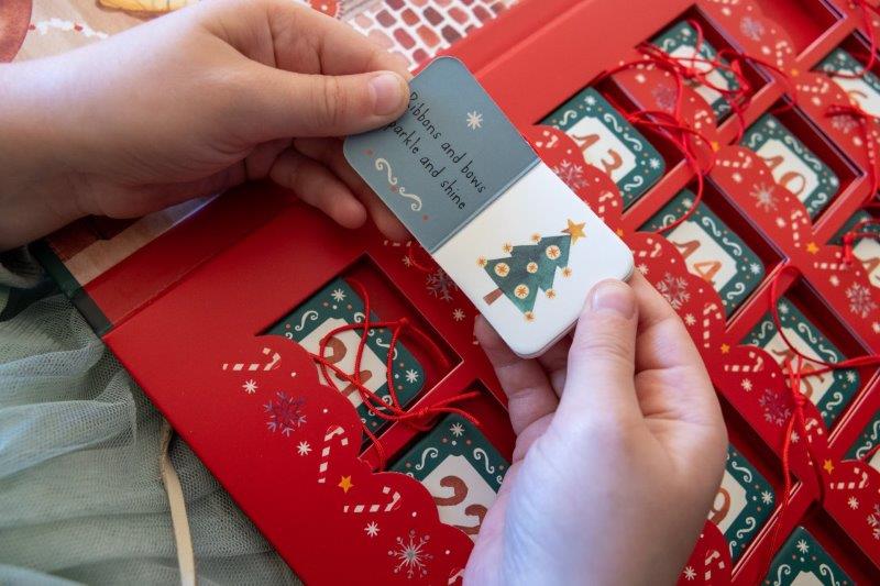 A person unveils the SASSI Christmas Advent Calendar, which features festive red packaging that perfectly embodies the holiday spirit. Inside, there is a card adorned with a heartfelt message and an illustration of a charming Christmas tree. Similar cards are visible, elegantly tied with red strings, each one adding a touch of magic to every day of the Christmas season.