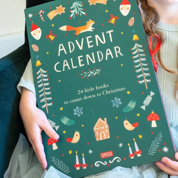 A person holds a Christmas Advent Calendar by SASSI, exuding holiday spirit with its festive illustrations of a fox, snowflakes, trees, and mushrooms. The cover announces: "Christmas Advent Calendar - 24 little illustrated books to count down to the Christmas season.