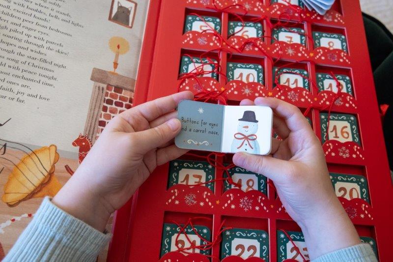 A child opens a SASSI Christmas Advent Calendar, revealing a card featuring a snowman illustration. The card reads, "Buttons for eyes and a carrot nose." Next to the calendar, an illustrated book captures the magic of the Christmas season with its festive artwork.