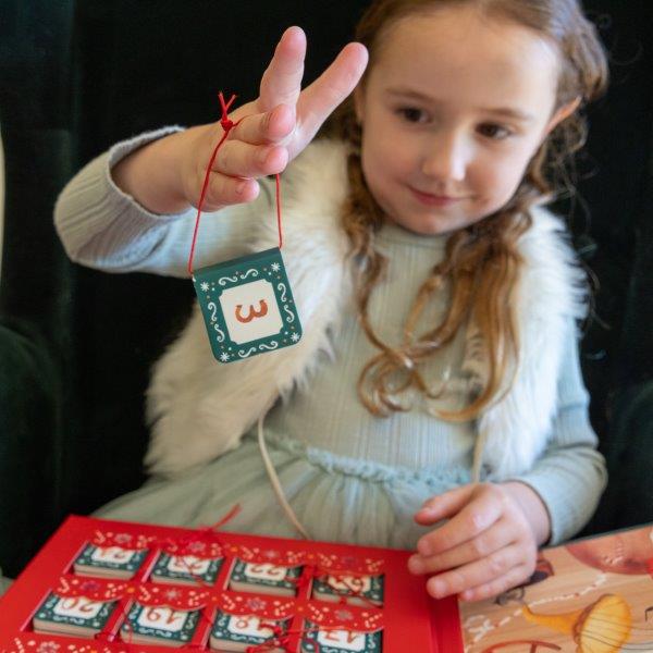 A young girl with long hair lifts a small ornament displaying the number three, embodying the holiday spirit. She sits before a SASSI Christmas Advent Calendar, while wearing a light blue dress paired with a white fur vest. The festive scene beautifully captures the warmth and joy of the Christmas season indoors.