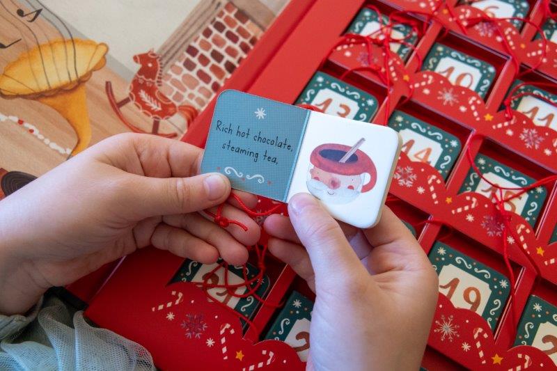 A person holds a small card showcasing a steaming cup of hot chocolate with the words "Rich hot chocolate, steaming tea." Celebrating the Christmas season, the card is placed next to SASSI's Christmas Advent Calendar, featuring red numbered compartments filled with festive items.