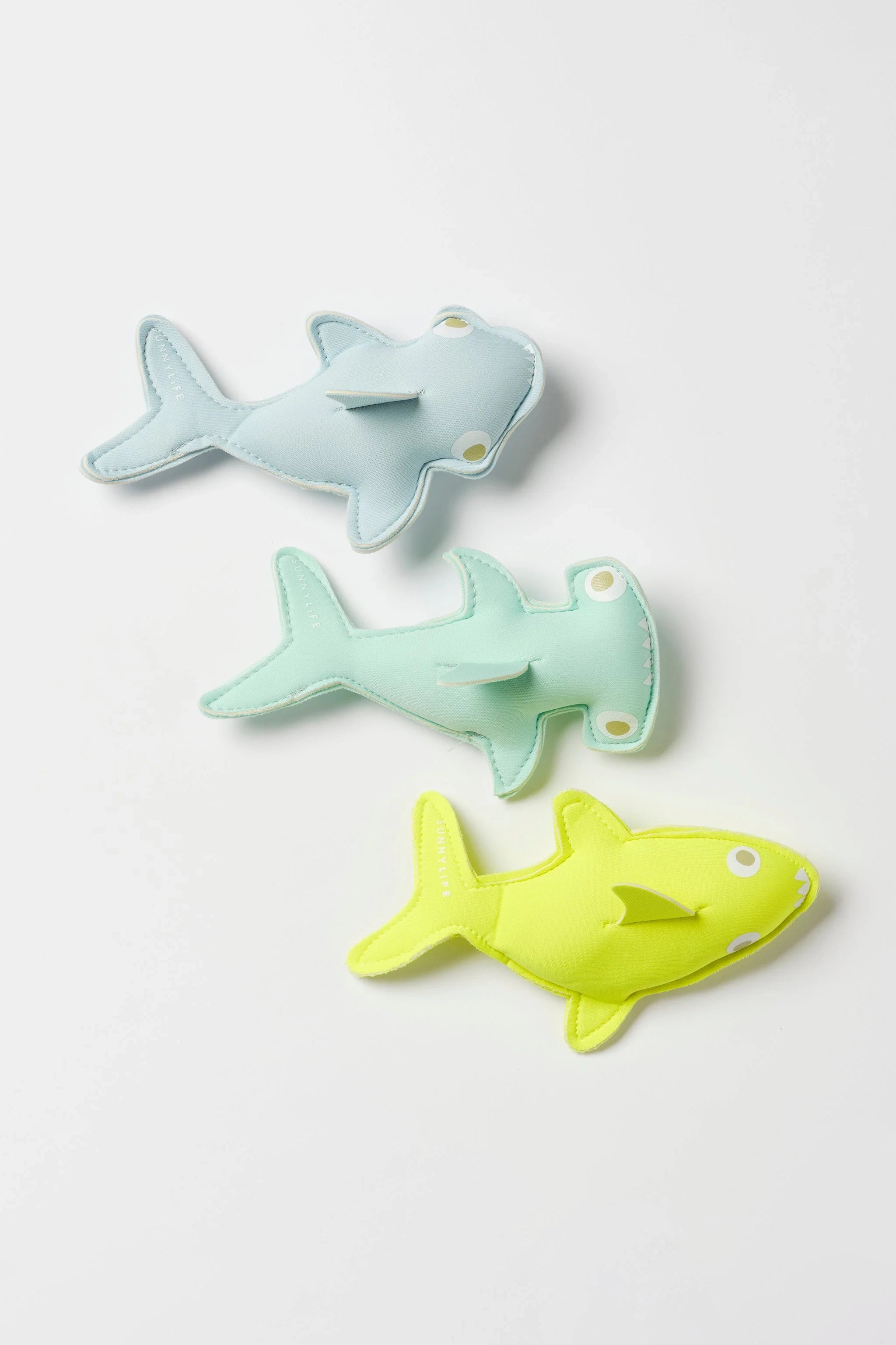 Three plush toy sharks from the SUNNYLIFE Salty the Shark Dive Buddies set are arranged on a white background. The top shark is light blue, the middle shark is light green, and the bottom shark is bright yellow. Made from durable neoprene, each shark features embroidered eyes and simple facial details, perfect for inspiring children’s diving adventures.