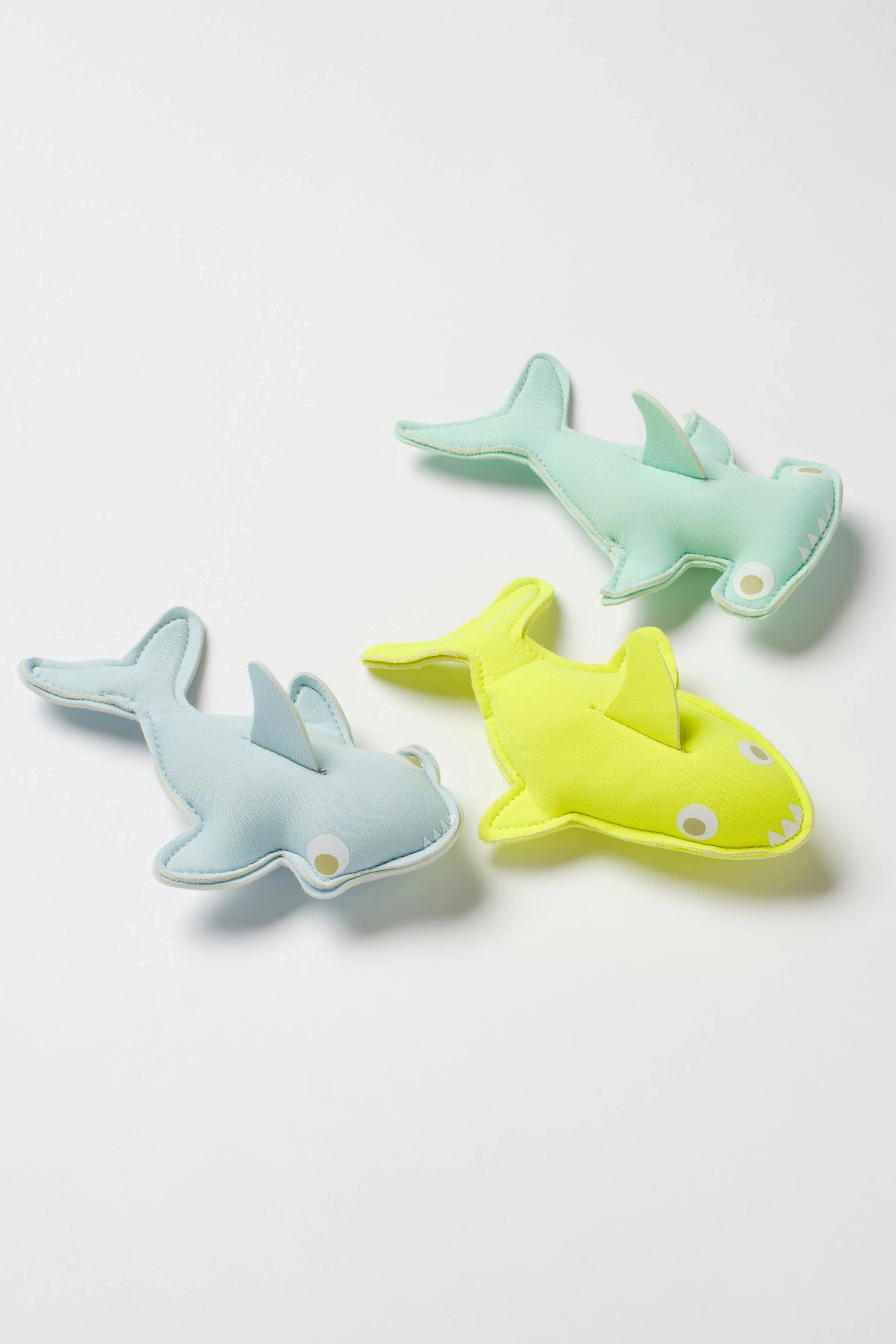 The Salty the Shark Dive Buddies (Set of 3) from SUNNYLIFE are displayed on a white surface. Each toy shark, made of durable neoprene fabric, comes in different vibrant colors: one in light blue, another in light green, and the third in bright yellow with a cheerful white smile and eyes—ideal for enhancing children's diving skills.