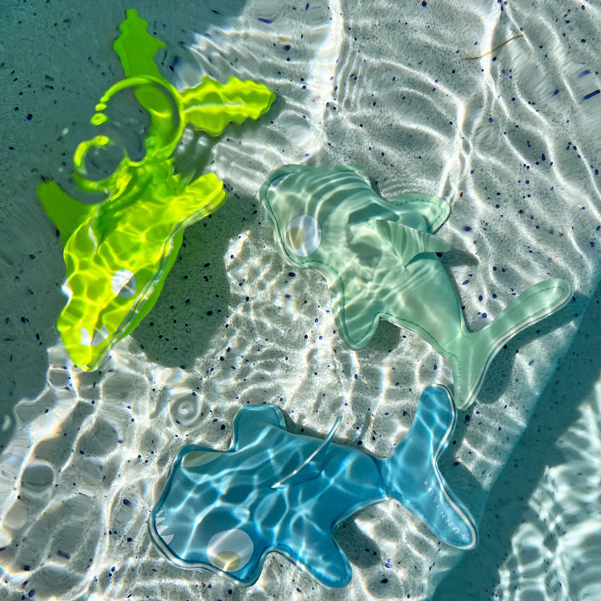 Three vibrant Salty the Shark Dive Buddies—one yellow, one green, and one blue—float in clear, sunlit water. Made by SUNNYLIFE from durable neoprene, they help improve children's diving skills as the water's ripples create dynamic reflections and shadows on the toys and the sandy floor beneath them.