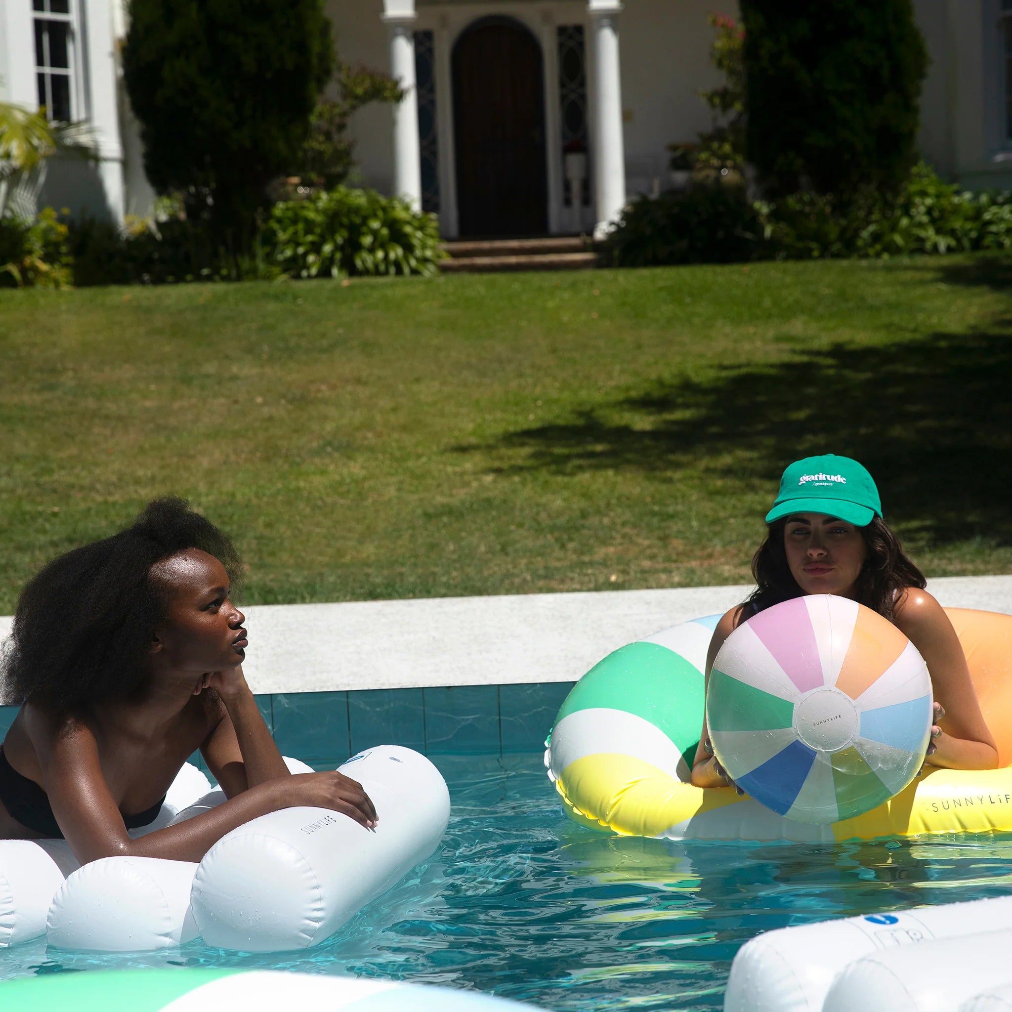 Two people are relaxing on inflatable pool floats in a swimming pool. One person is on a white float and the other is on a pastel-colored SUNNYLIFE float, holding an Inflatable Beach Ball Pastel Gelato by SUNNYLIFE. In the background, there are lush green plants and a white house with columns, creating an idyllic setting.