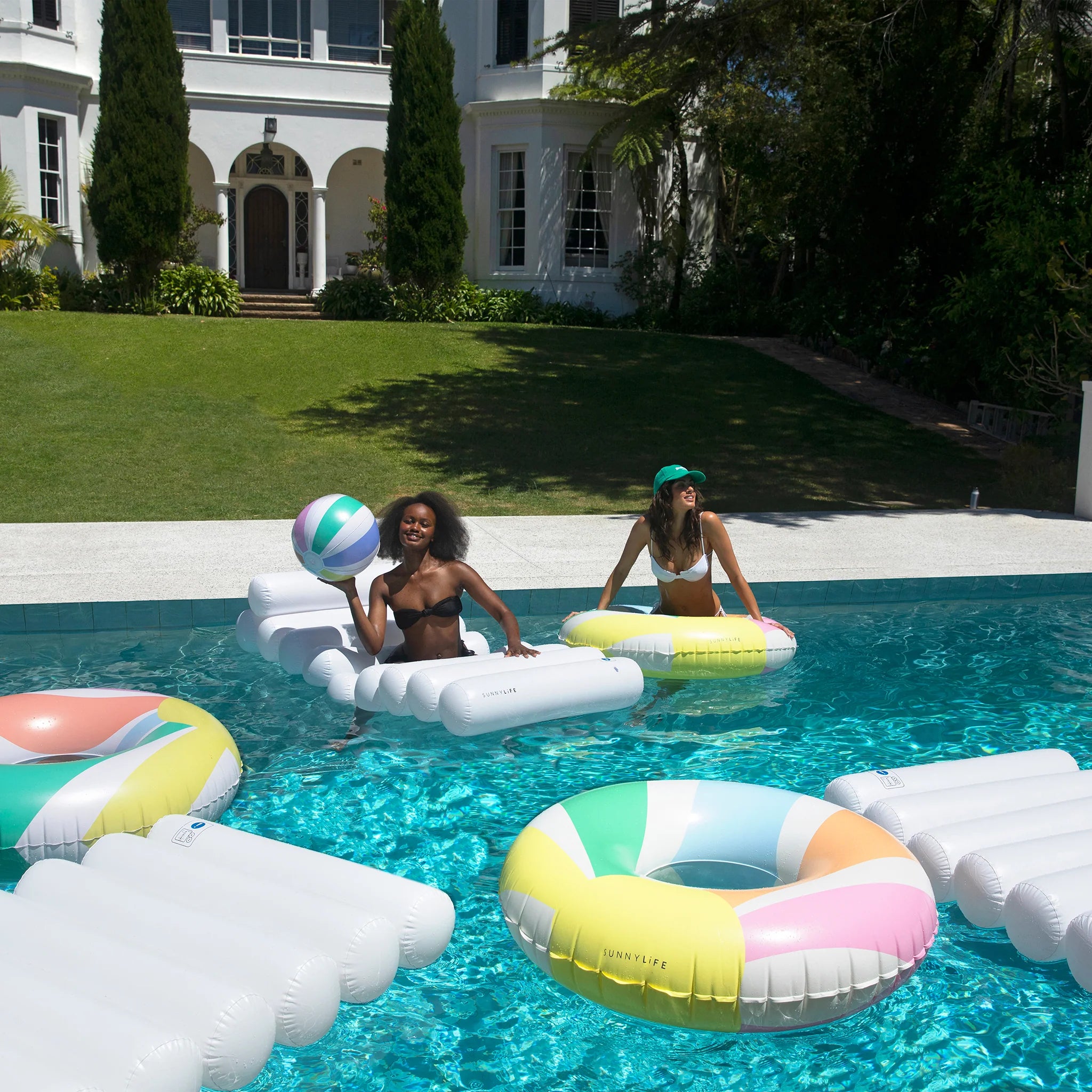 Two women enjoy a sunny day in a clear blue pool surrounded by colorful SUNNYLIFE floats. One woman, standing on a raft, holds an Inflatable Beach Ball Pastel Gelato by SUNNYLIFE. The other woman, seated on a donut-shaped float made of durable PVC, wears a mint green cap. A large, white house and manicured lawn are in the background.