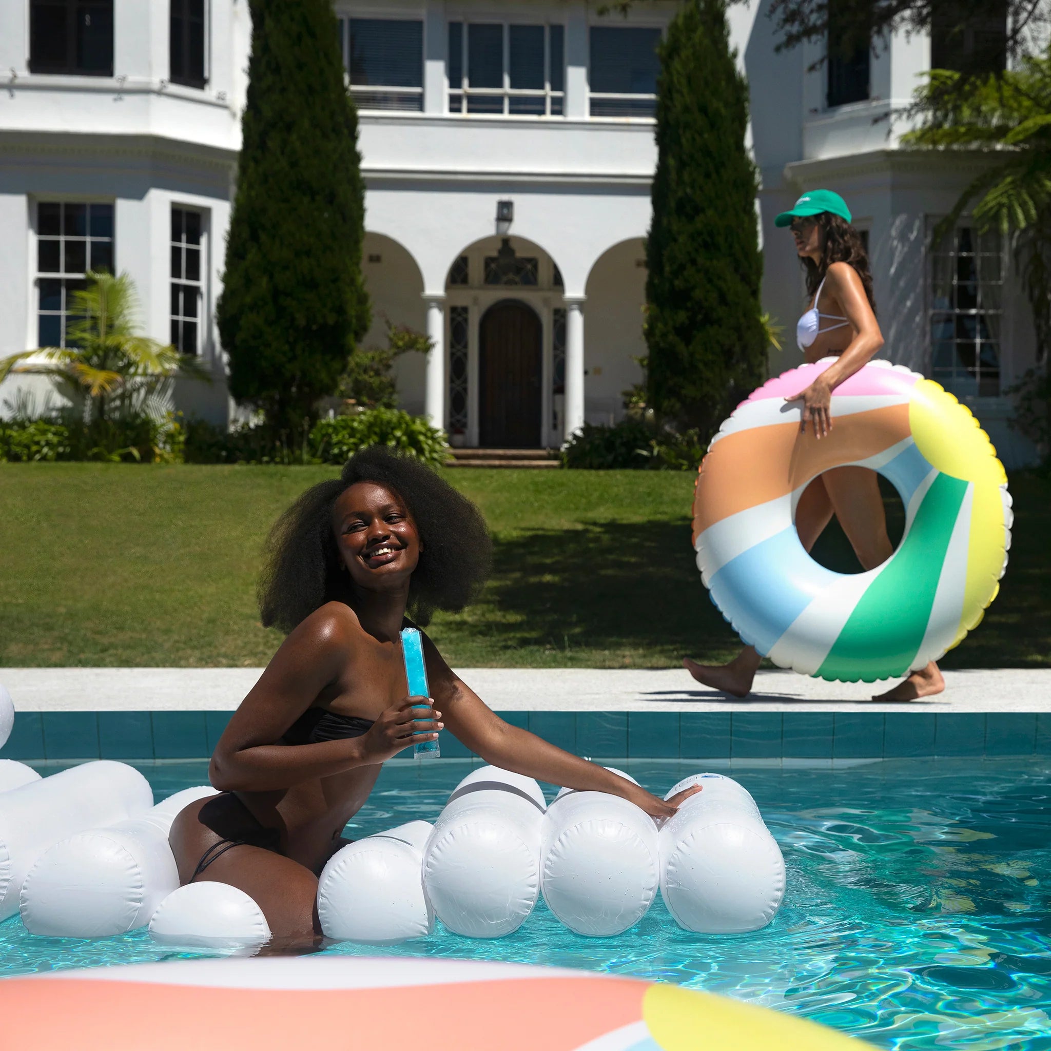 A woman smiles while sitting on a durable PVC, phthalate-free SUNNYLIFE Tube Float Pastel Gelato and holding a popsicle in a pool. In the background, another person walks on the lawn holding a colorful inflatable ring. A large white house with tall trees is visible behind them.