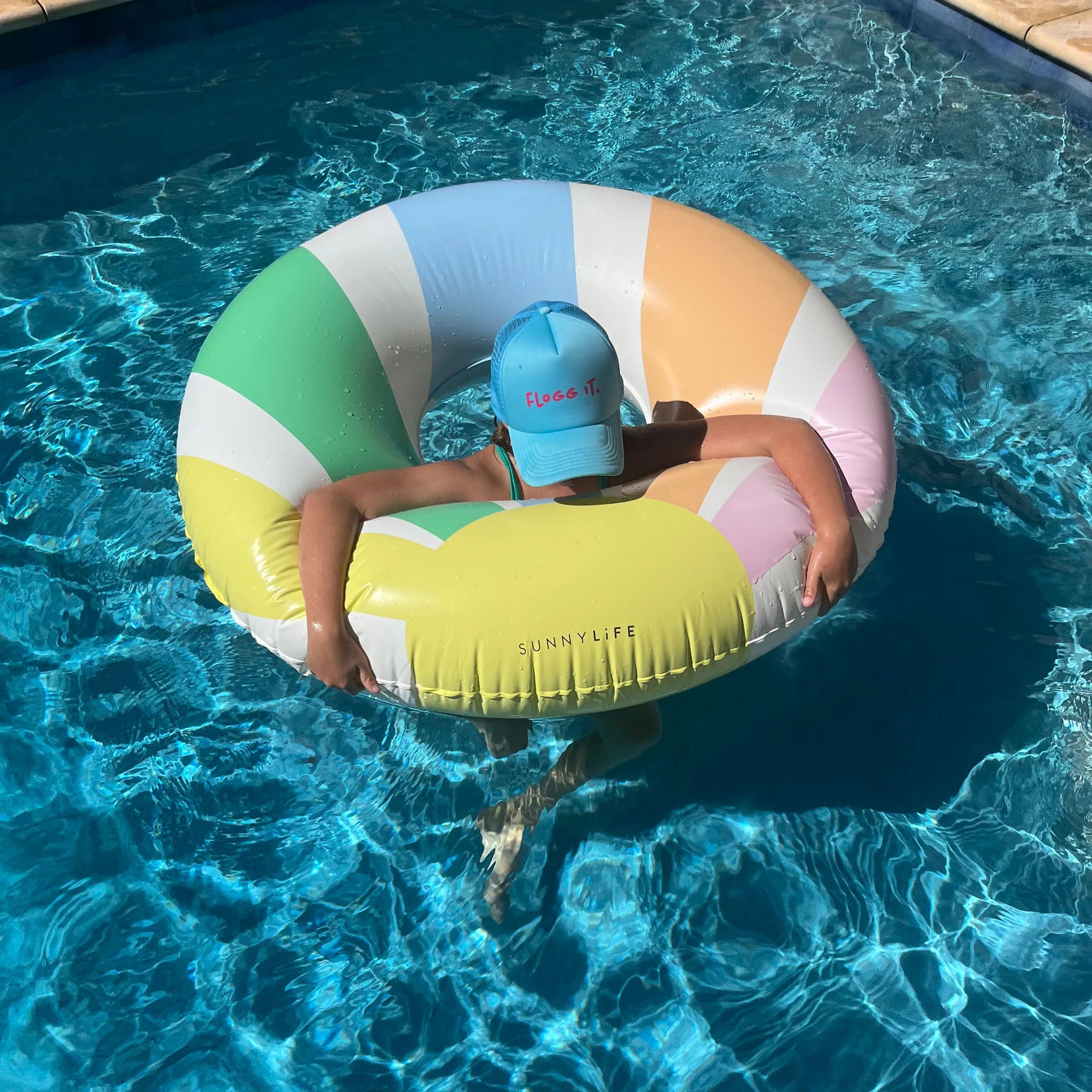 A child wearing a blue cap floats in a swimming pool, seated inside the large Sunnylife Tube Float Pastel Gelato, made of durable PVC. The bright sun shines on the clear blue water, creating a lively and cheerful summer atmosphere with their phthalate-free SUNNYLIFE float.