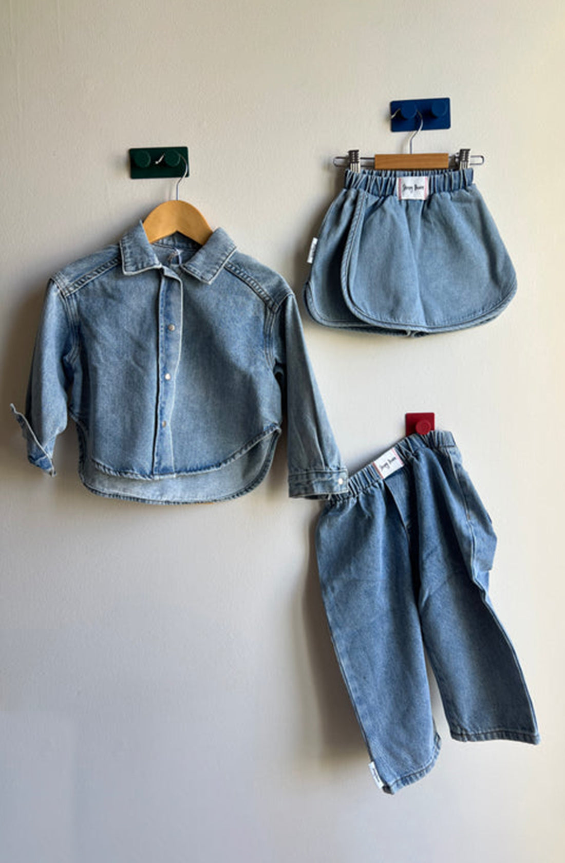 A children's denim outfit from SLEEPY DENIM is displayed against a light-colored wall. It includes a long-sleeve denim button-up shirt (left), a denim skirt (top-right), and a versatile piece—a Dawning Shacket Sky Blue Wash (bottom-right). Each item, crafted from organic cotton, is hung by colored clips: green, blue, and red.