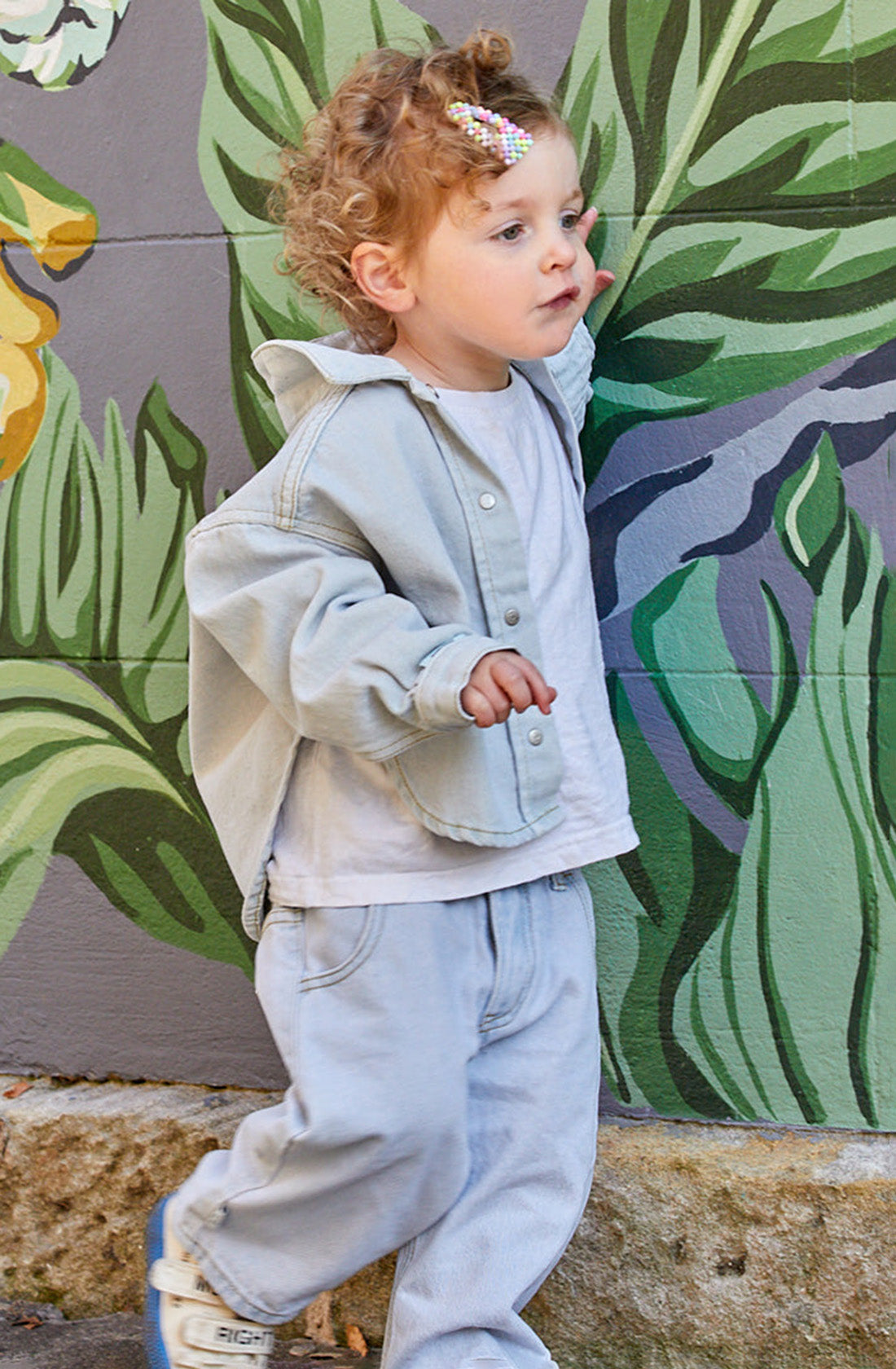 A toddler with curly hair, adorned with a colorful hair clip, is dressed in the "Dawning Shacket Cloud Wash" from SLEEPY DENIM, alongside a white shirt and light jeans. They are walking beside a mural of large green leaves, touching the mural with one hand while looking off to the side.