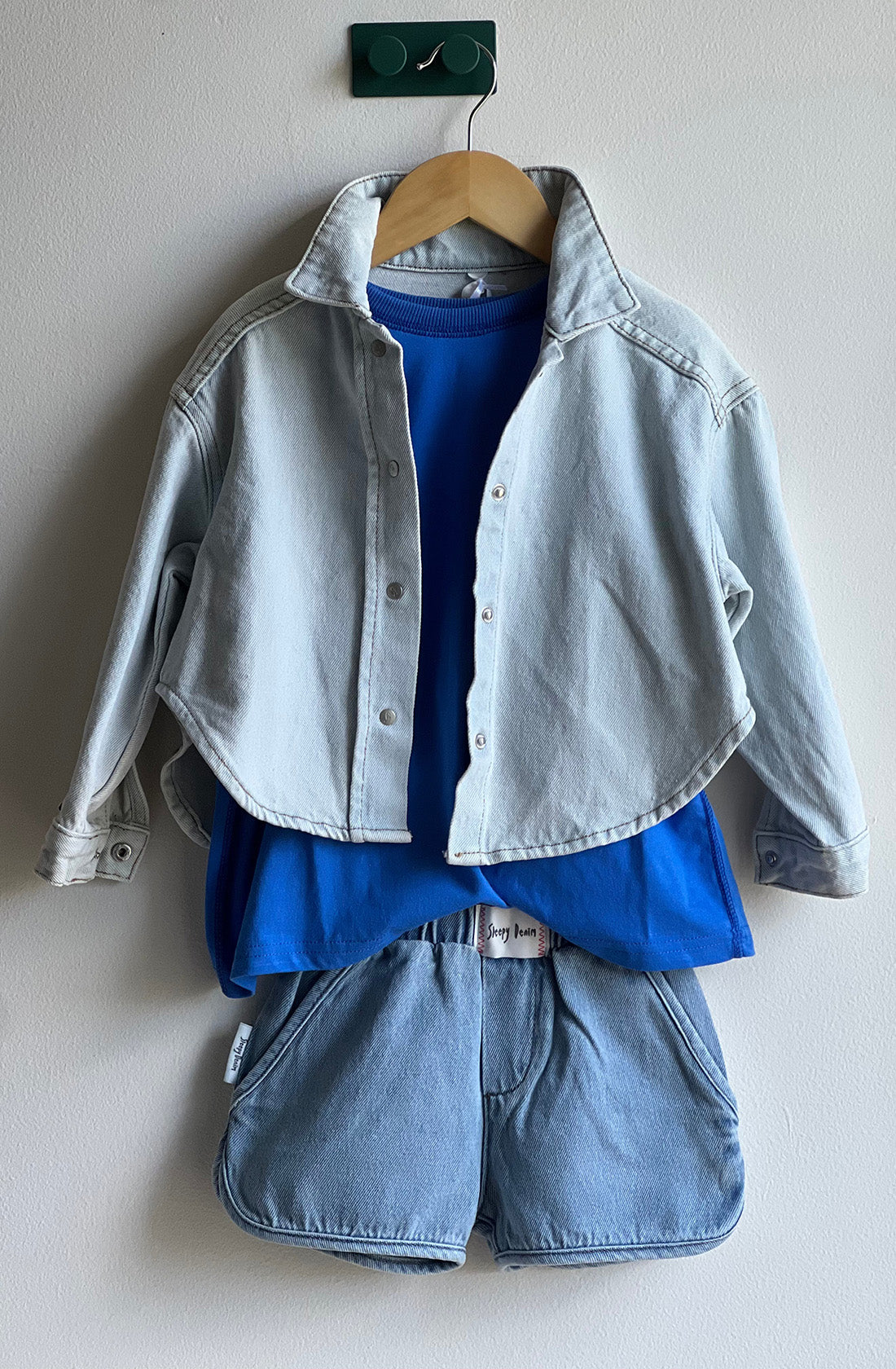 Displayed against a white wall is a children's outfit consisting of the Dawning Shacket Cloud Wash by SLEEPY DENIM, layered over a bright blue T-shirt, and paired with matching light blue denim shorts. This versatile wardrobe staple hangs neatly on a wooden hanger with a green hook attached to the wall.