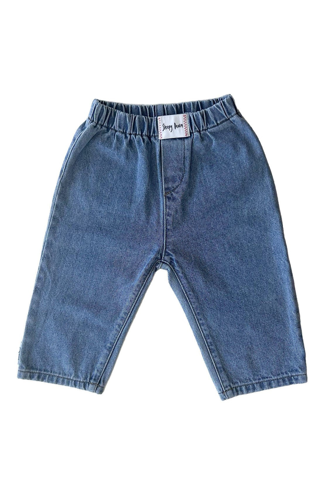 A pair of Raver Jean Sky Blue Denim jeans from SLEEPY DENIM, designed for infants or toddlers. Made from organic cotton, these jeans feature an elastic waistband and a loose fit. A small white tag with text is visible at the center of the waistband, set against a crisp white background.