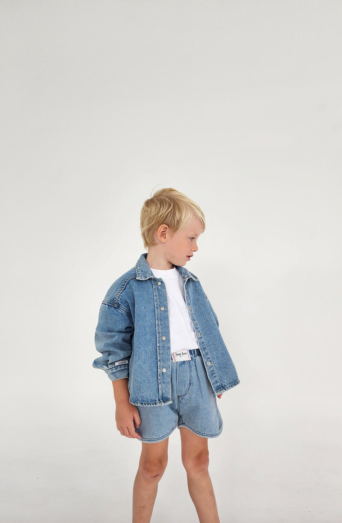 A young boy with blonde hair stands against a plain background, showcasing the Dawning Shacket Sky Blue Wash from SLEEPY DENIM. Paired with denim shorts and a white T-shirt, this versatile piece, made of organic cotton, is perfect for any casual outing. He looks to his right with his hands by his sides.