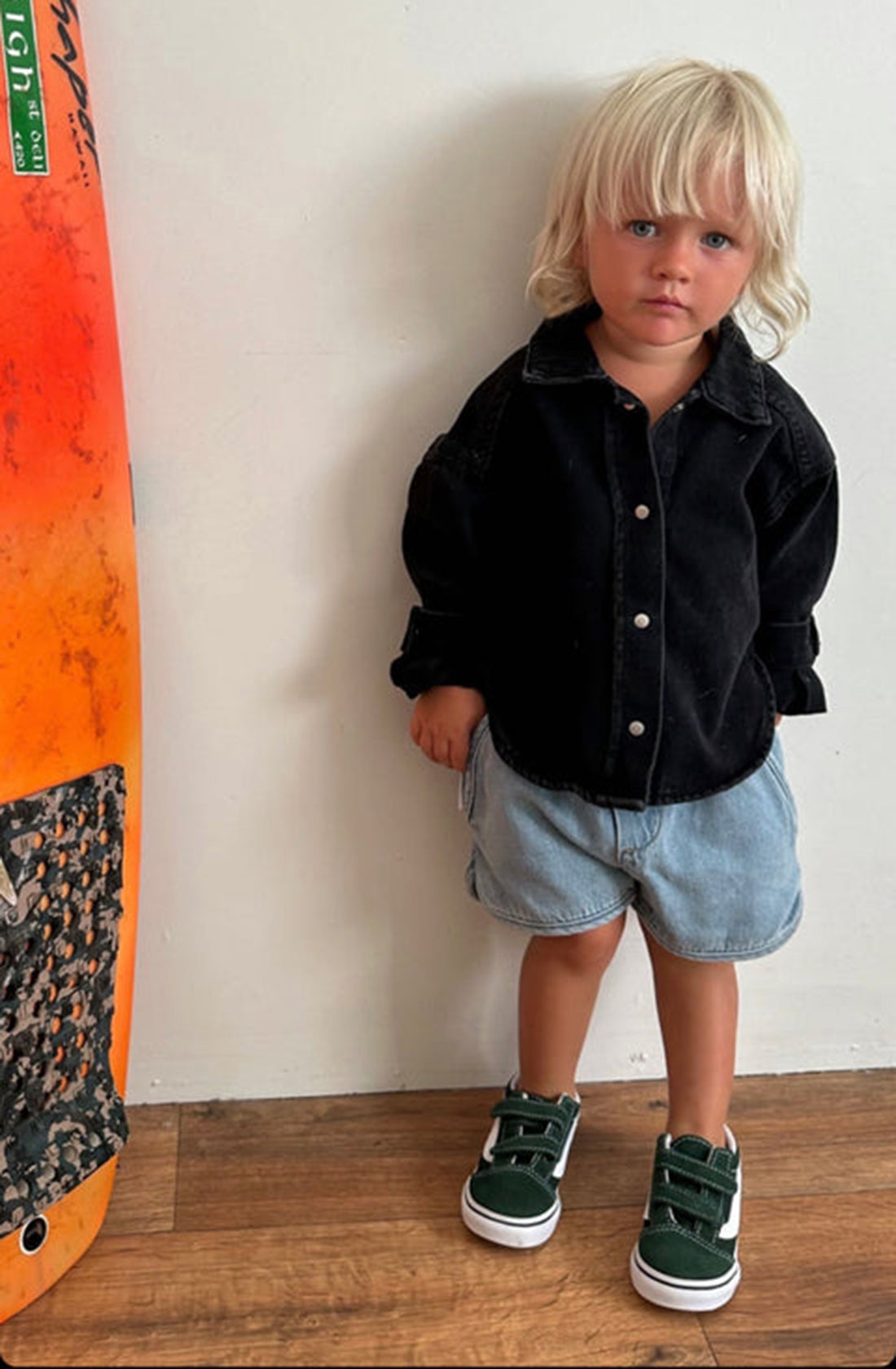 A young child with blond hair stands indoors, wearing a SLEEPY DENIM jacket, light blue Kip Shorts Sky Blue, and green velcro sneakers. An orange and black surfboard is propped against the wall beside them on the wooden floor. The child looks at the camera.