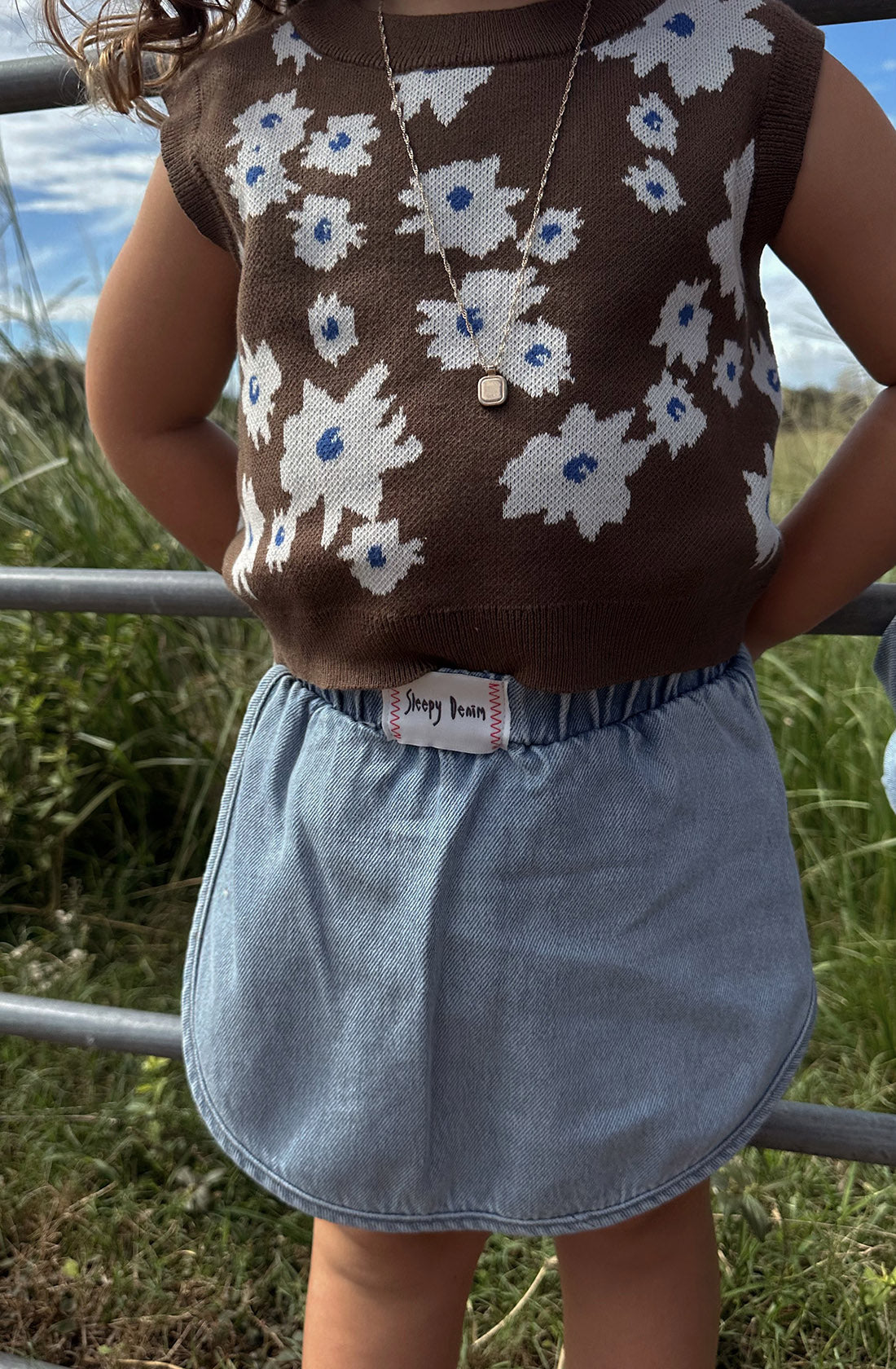 A child stands outdoors near a metal fence wearing a sleeveless brown top with a blue and white floral pattern, a necklace with a small pendant, and an organic cotton denim skort called the "Siesta Skort Sky Blue" from SLEEPY DENIM, featuring an elastic waistband. Grass is visible in the background.