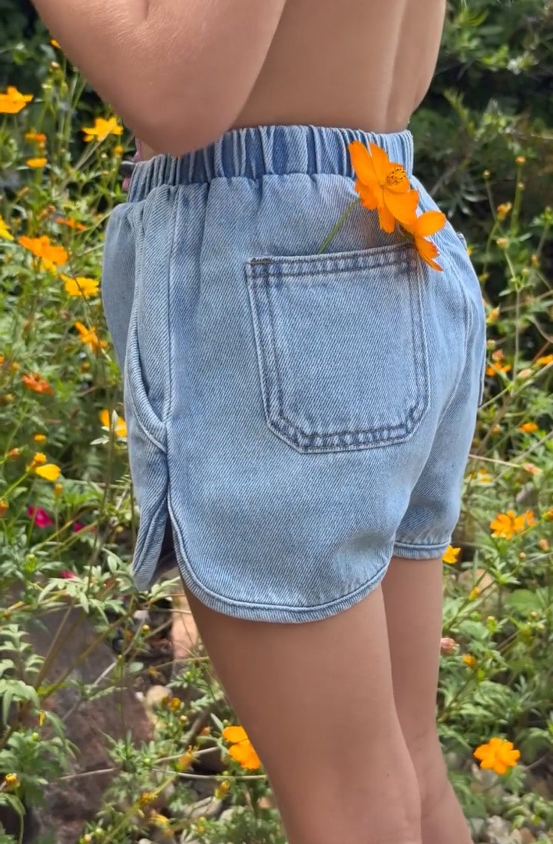 A person wearing organic cotton denim shorts, known as the Siesta Skort Sky Blue by SLEEPY DENIM, stands with their back to the camera. A vibrant orange flower is tucked into the waistband pocket. The background features lush greenery and several yellow and orange flowers in full bloom.