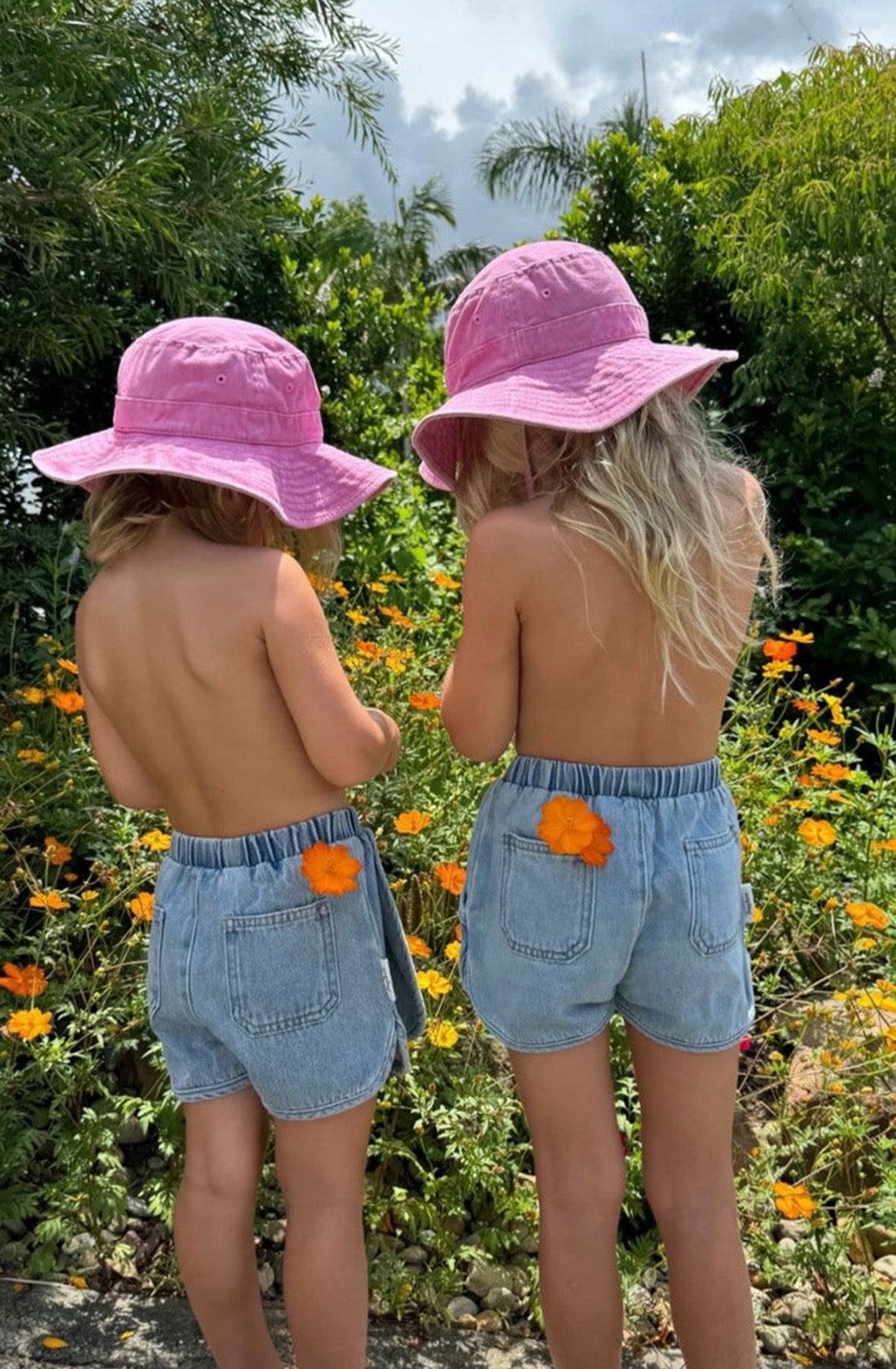 Two young children with long blonde hair, wearing pink sun hats and SLEEPY DENIM's Siesta Skort in sky blue, stand with their backs to the camera in a garden filled with greenery and orange flowers. An orange flower is tucked into each of their back pockets.