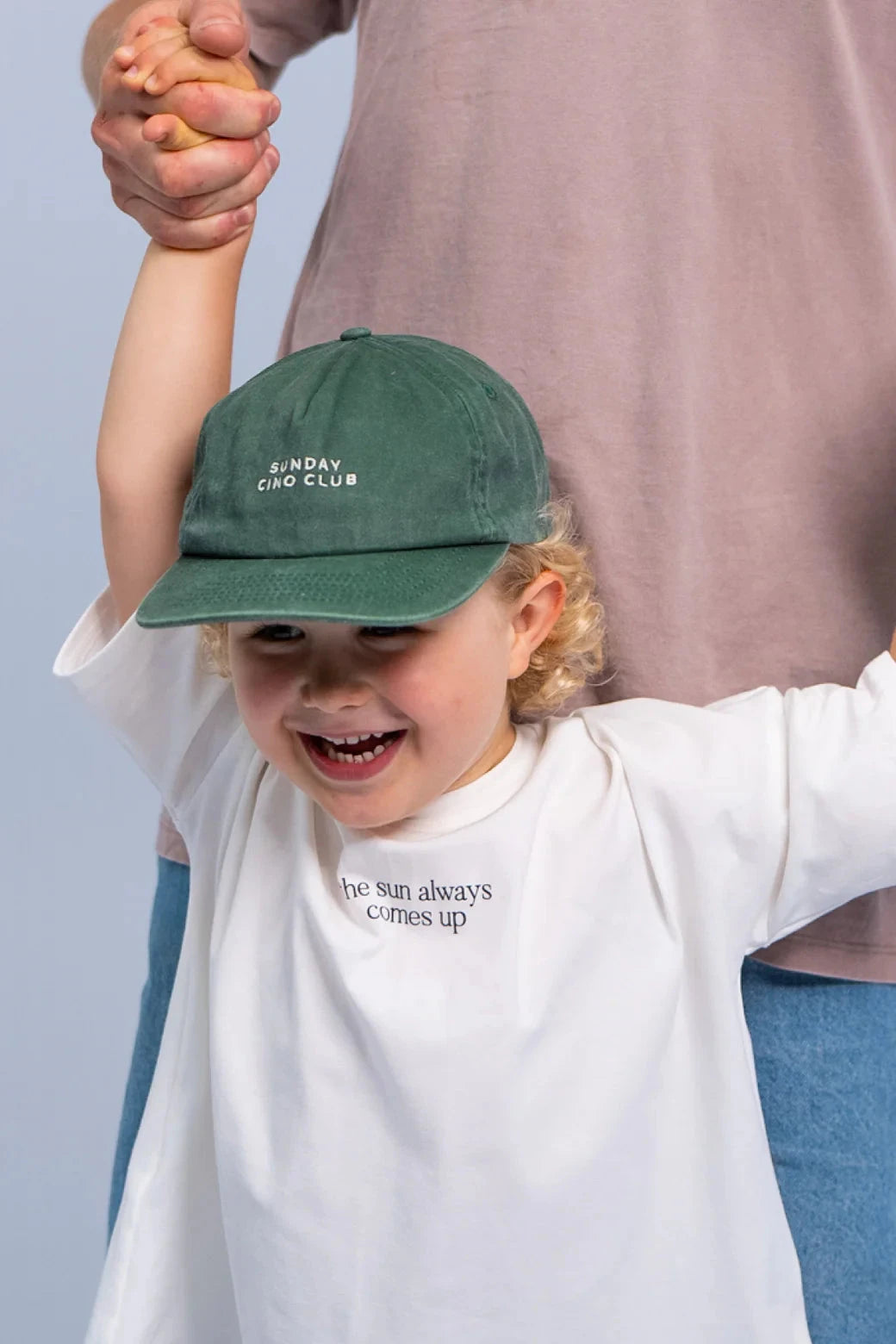 A young child wearing a SONNY LABEL Sunday Cino Club Cap in Basil, featuring an adjustable brass strap with "SUNDAY CINO CLUB" embroidered on it, paired with a white shirt that reads "the sun always comes up." The child is smiling and holding up their hands, possibly supported by an adult from behind.