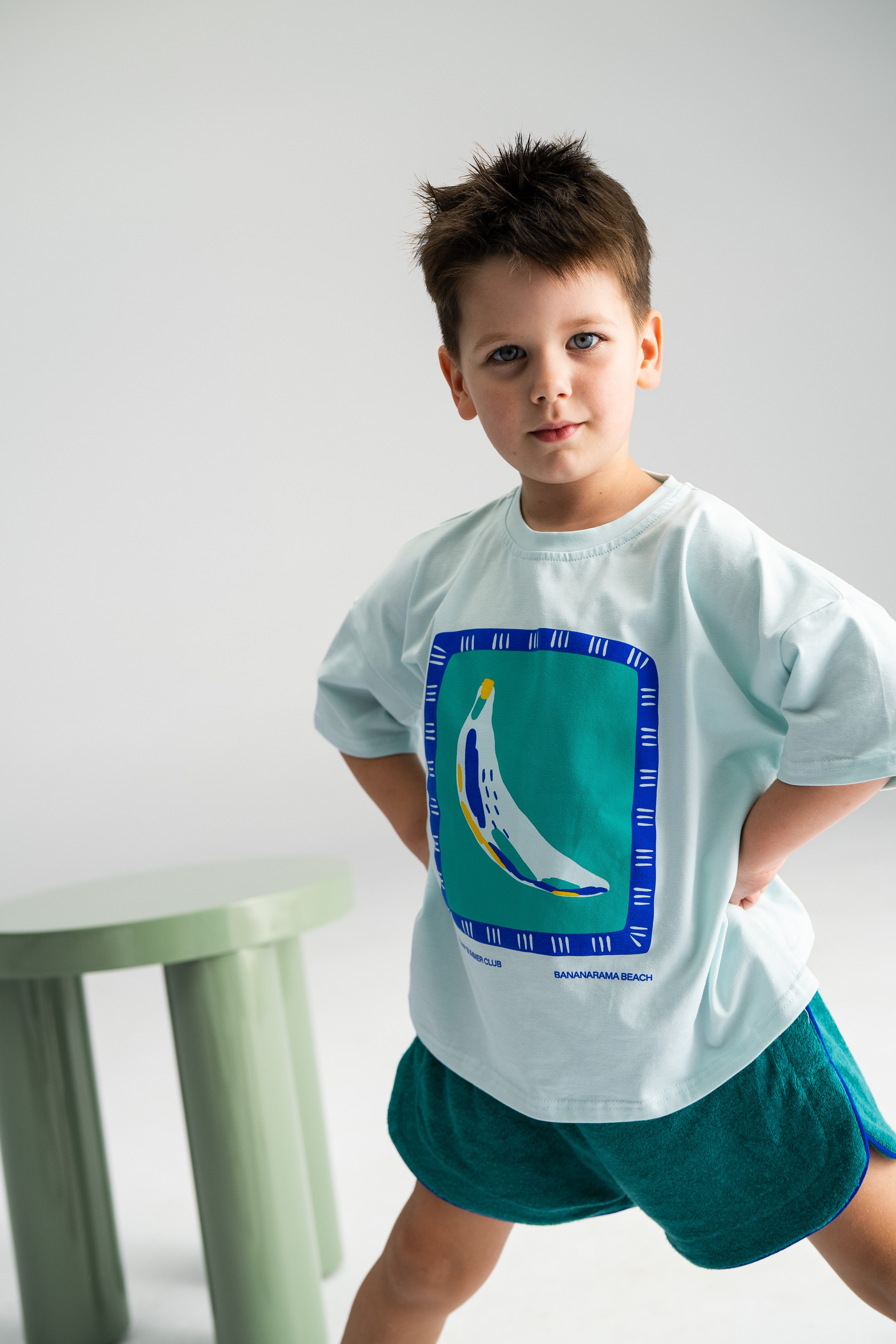 A young boy with short hair stands confidently with his hands on his hips, wearing the SONNY LABEL's signature Bananarama Tee Baby Blue, featuring an abstract banana design and teal shorts. Near him, a light green stool rests on a plain background, capturing the essence of Bananarama Beach style.