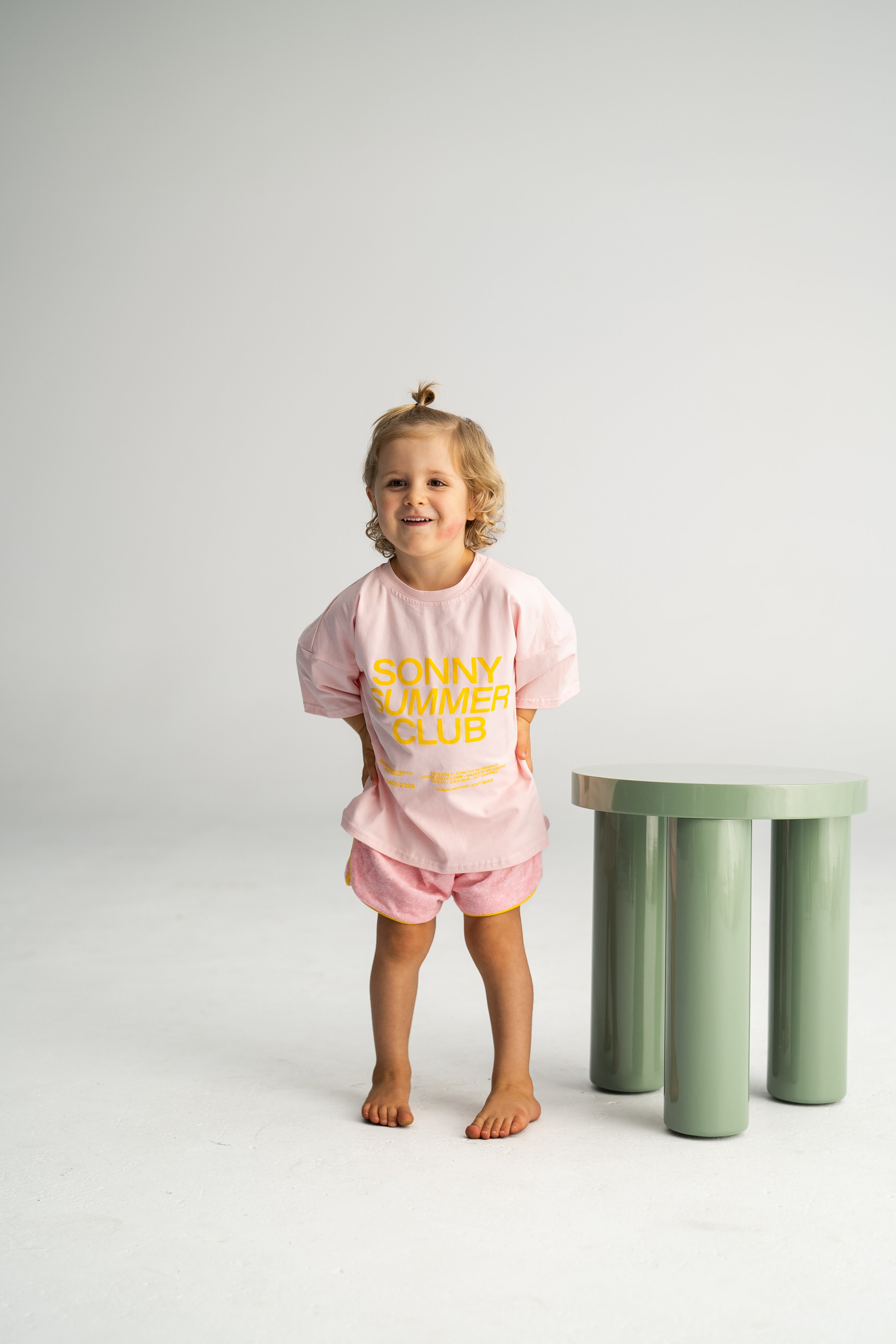 A young child with light curly hair stands barefoot on a plain white background, smiling. Dressed in a *Summer Club Tee Pink* by SONNY LABEL and shorts, they evoke the carefree spirit of the Sonny Summer Club, standing next to a small, green, cylindrical stool.