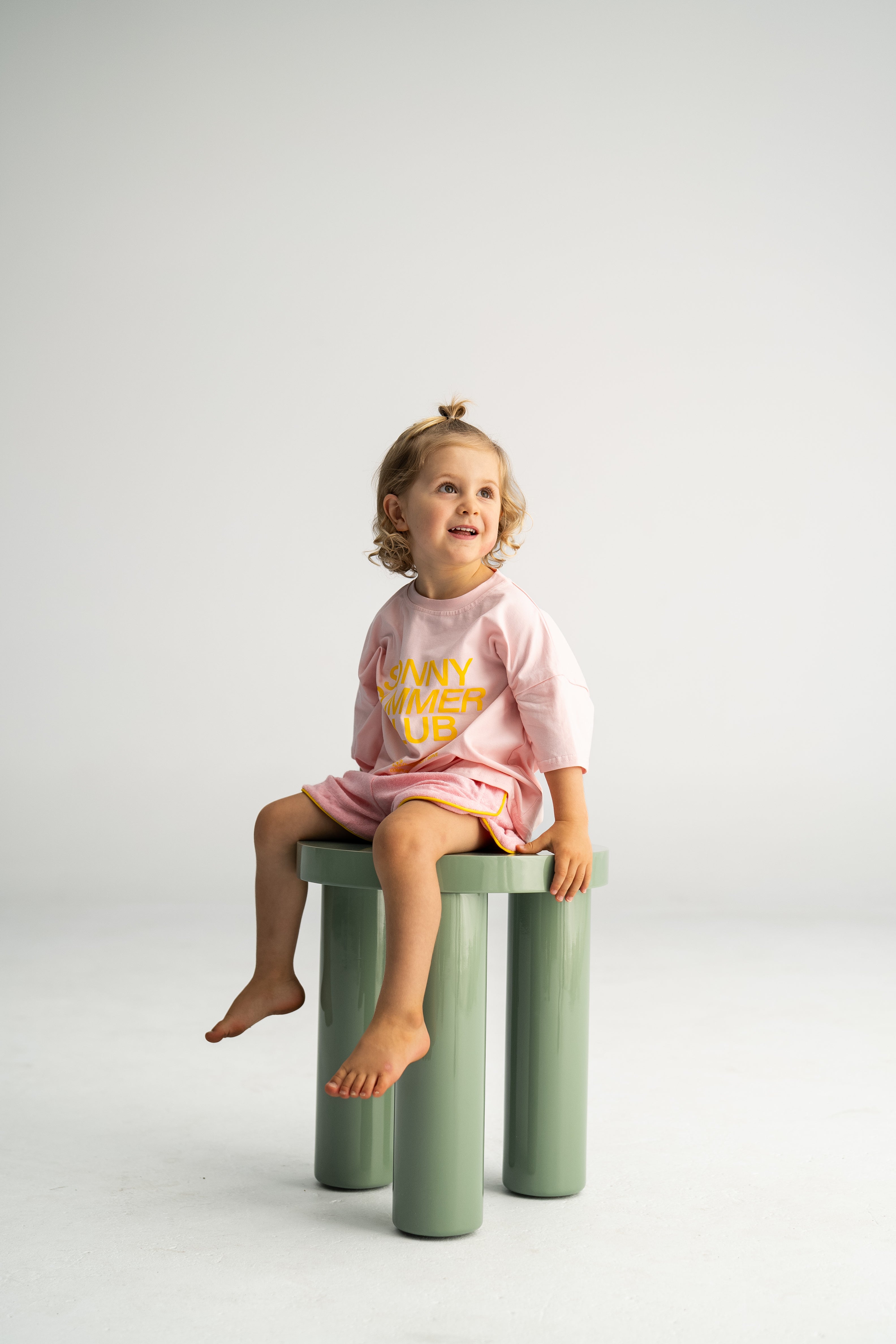 A small child with curly hair sits barefoot on a green, cylindrical stool, dressed in the Terry Shorties Pink by SONNY LABEL. The light pink outfit is adorned with yellow text and has a soft cotton terry texture. The backdrop is a simple, light gray.