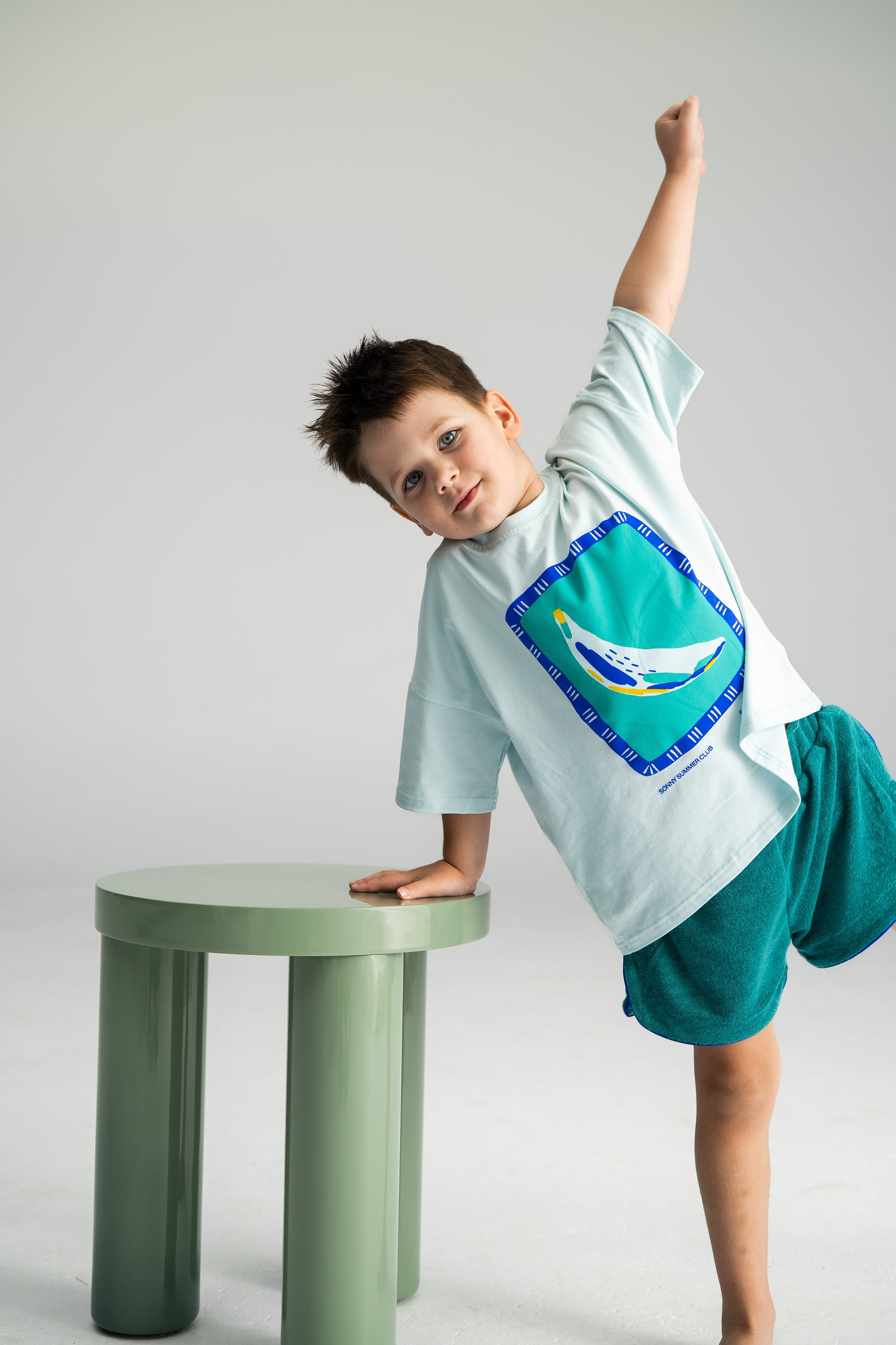 A young boy with short hair leans on a green table, his right arm raised triumphantly. He wears a light blue t-shirt with a bird graphic and Terry Shorties Teal by SONNY LABEL, standing on one leg in an airy, minimalist setting.