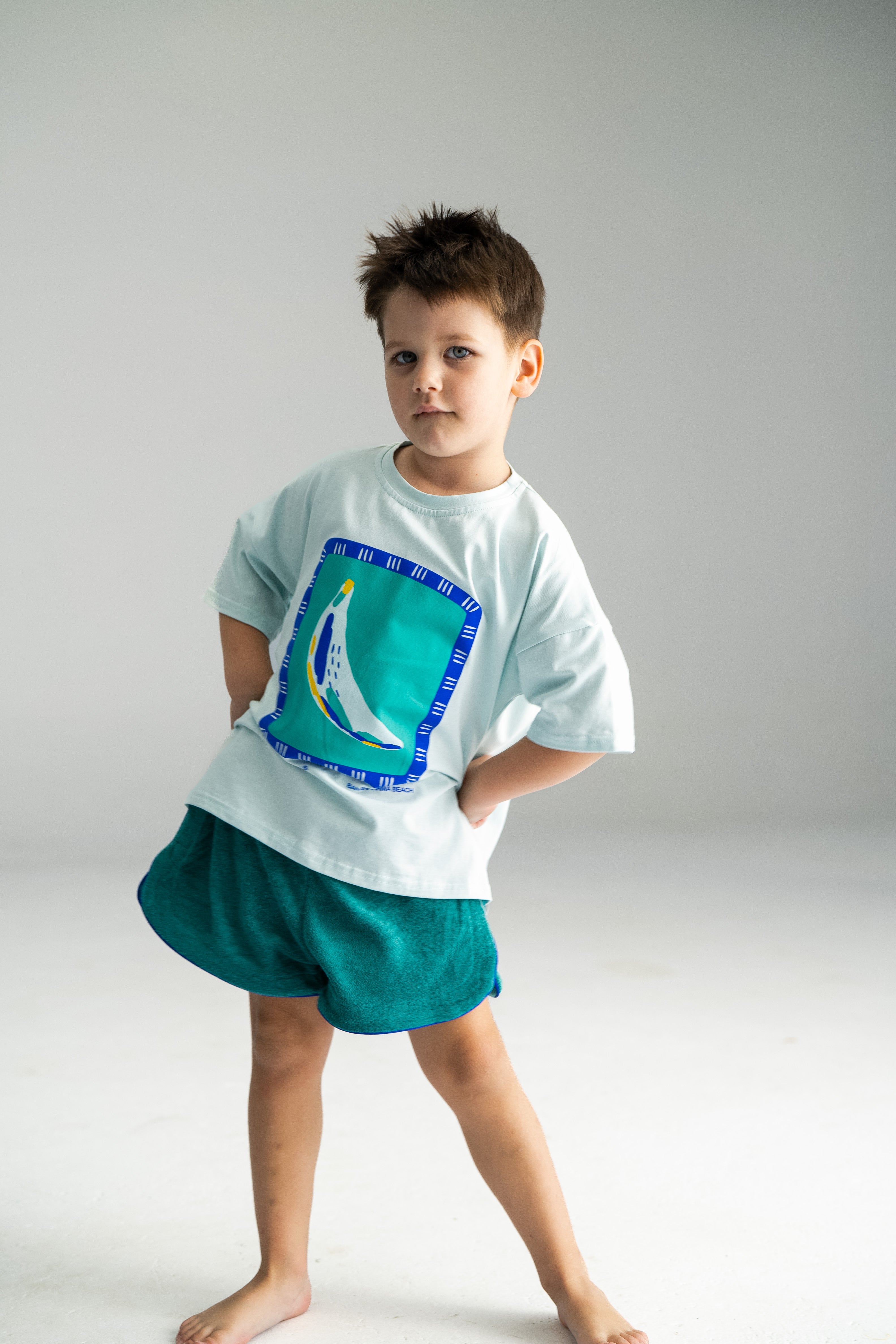 A young child stands confidently on a plain background, wearing a light blue cotton terry shirt adorned with a colorful shoe graphic and pairing it with SONNY LABEL's Terry Shorties Teal. With a playful expression and slightly tousled hairstyle, the child is barefoot, exuding joy and spontaneity.