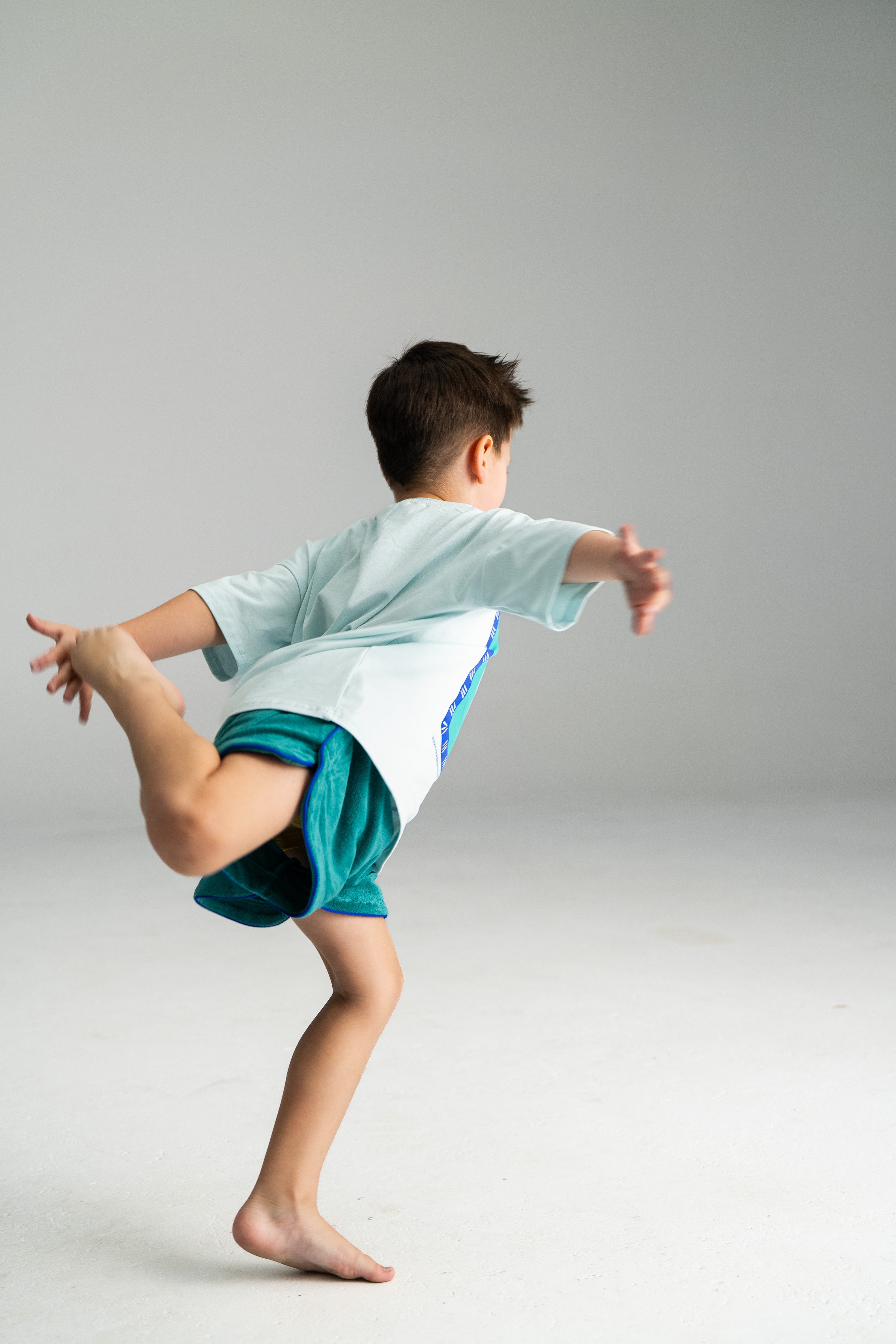 A child clad in a light blue shirt and SONNY LABEL's Terry Shorties in teal balances gracefully on one leg, bending the other behind and holding it with their hand. The loose fit of the shorts allows for freedom of movement as they stand barefoot on a plain white surface, striking an elegant pose against a simple gray background.