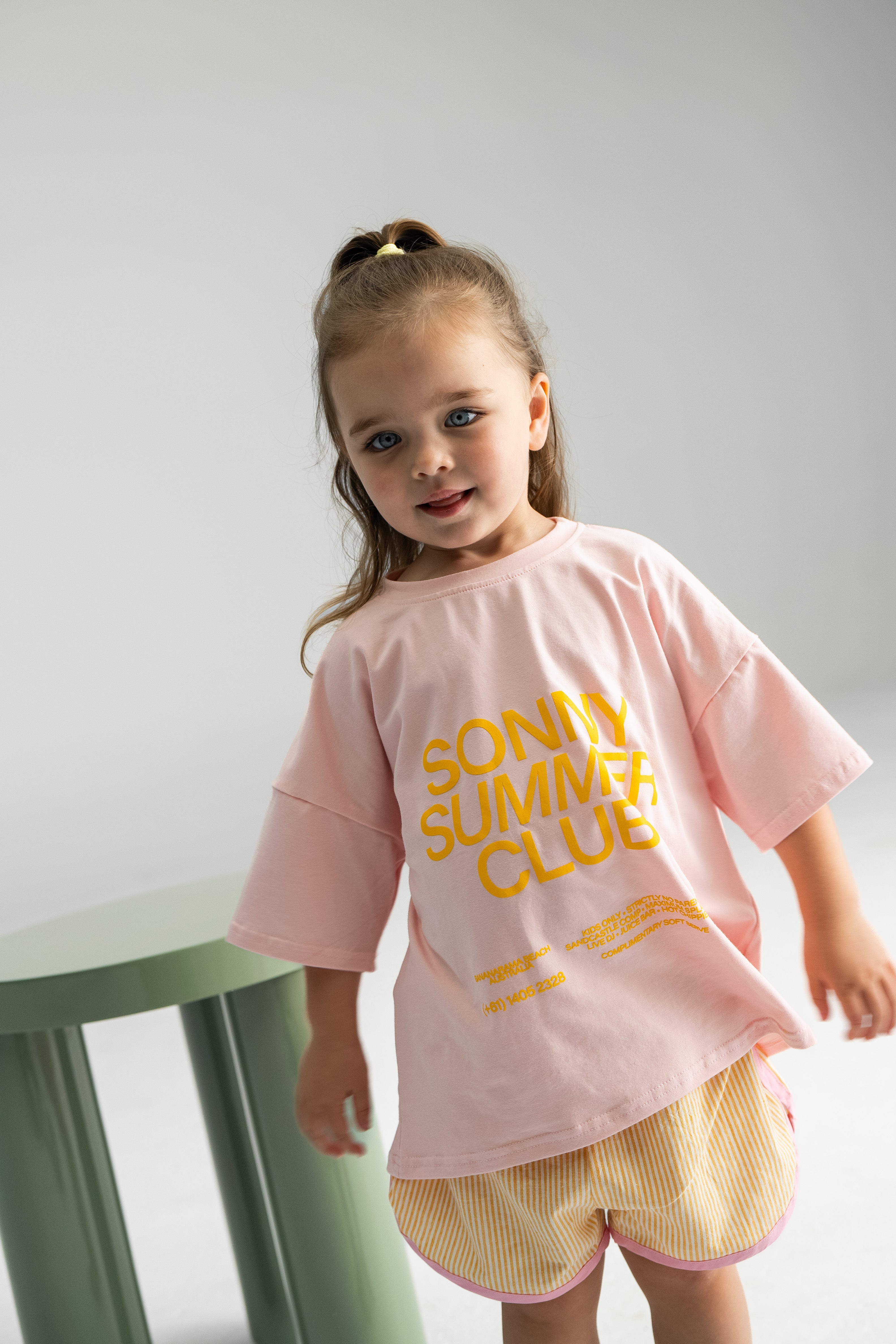 A young child with long hair tied up wears a light pink T-shirt featuring yellow text that reads "Sunny Summer Club" and the Stripe Shorties Lemon by SONNY LABEL. They are indoors beside a light green stool, offering a slight smile while enjoying the summer comfort against a neutral background.