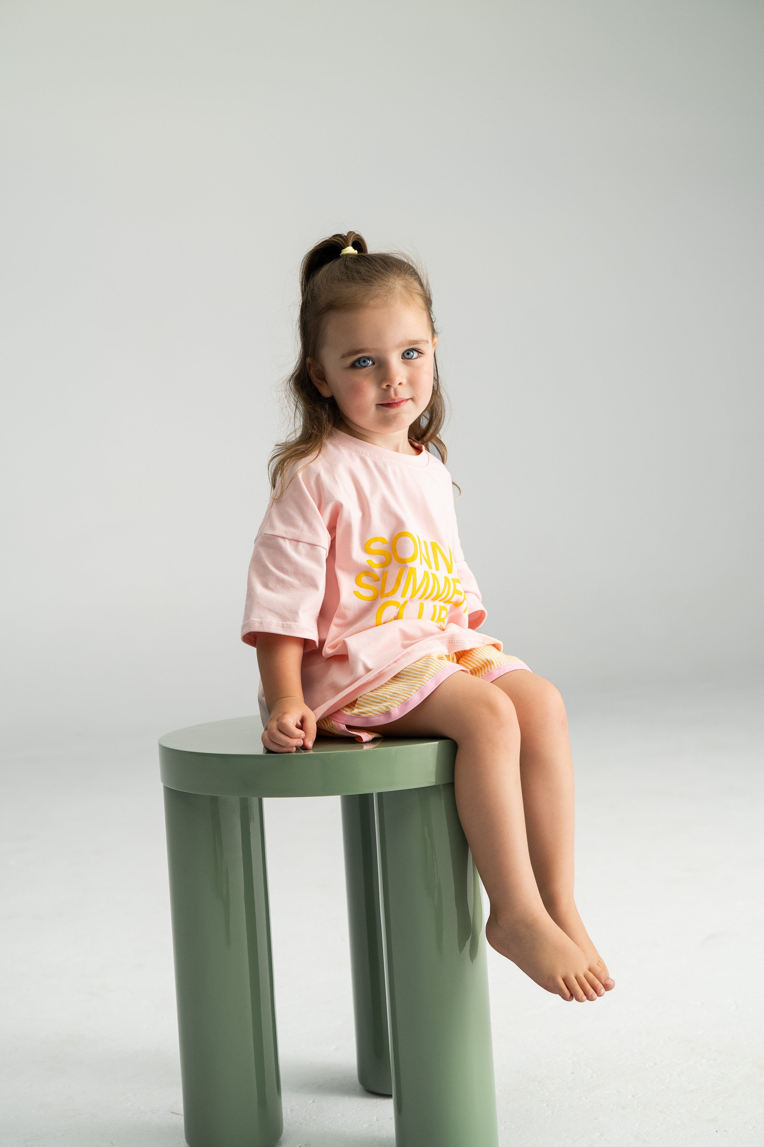 A young girl with long hair sits on a vibrant green stool, wearing a pink T-shirt featuring eye-catching yellow text and contrasting pink edging. She holds a pair of stylish Stripe Shorties Lemon from SONNY LABEL, perfectly capturing the essence of summer comfort against a soft and minimalistic backdrop.