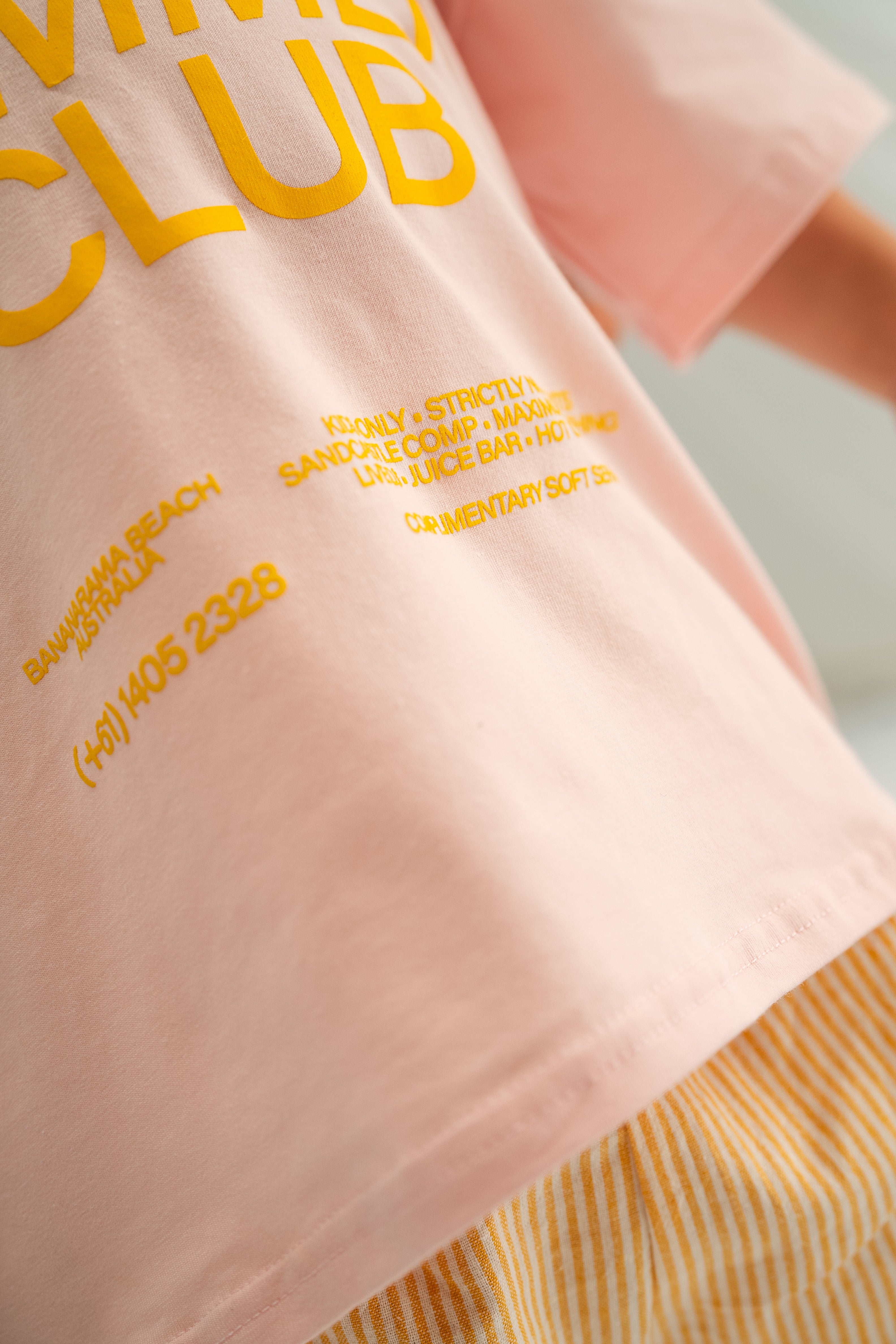 A close-up view shows someone wearing the Summer Club Tee Pink by SONNY LABEL, featuring yellow text with "Sonny Summer Club" and a phone number. This fun tee perfectly complements striped yellow and white pants, capturing the lively essence of Bananarama Beach.