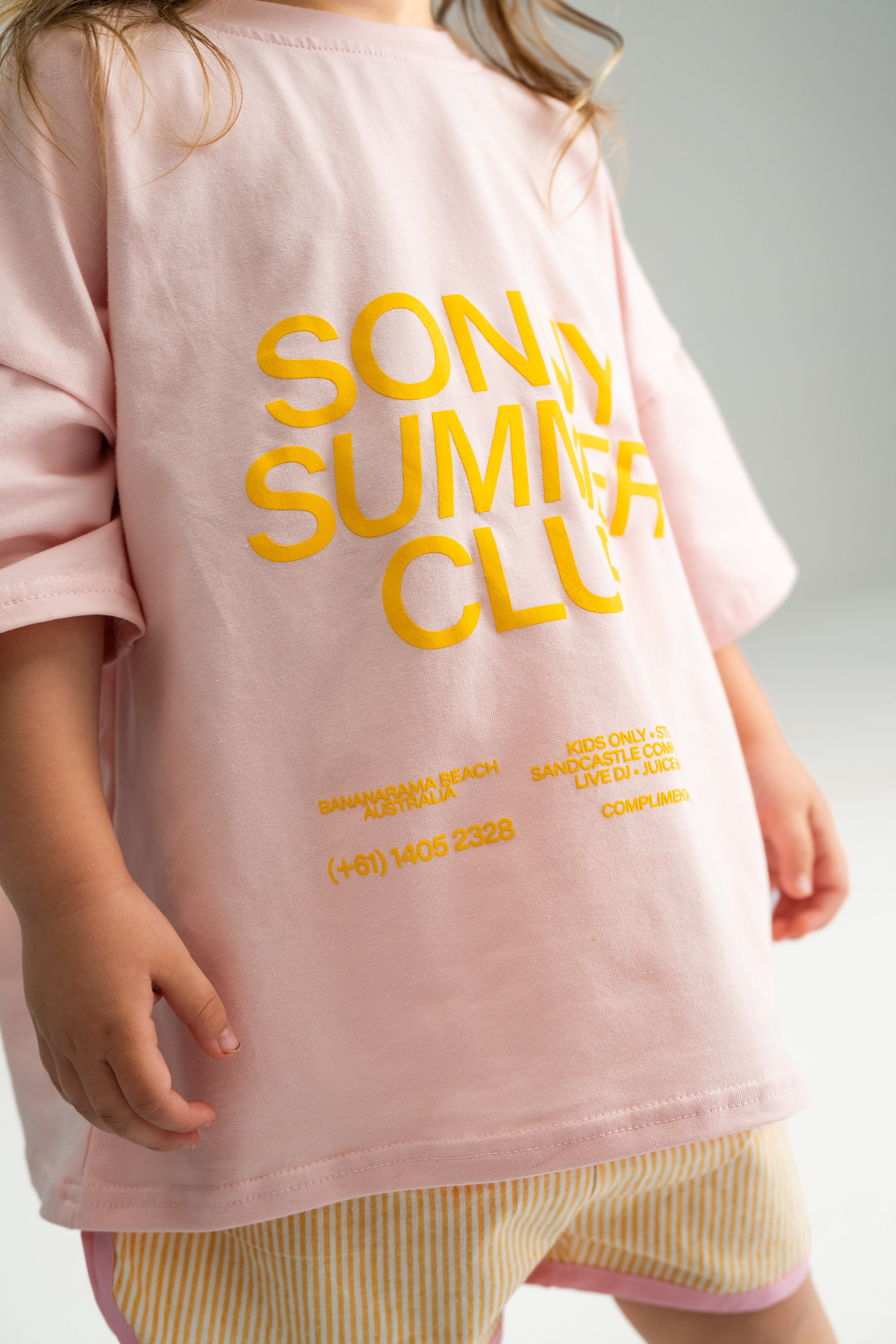 A child wearing a light pink Summer Club Tee by SONNY LABEL, featuring yellow text that reads "Sonny Summer Club," along with striped shorts, stands against a light background. The tee also includes small text with a phone number and other details, emphasizing this is exclusive kids' fun at the lively Bananarama Beach.