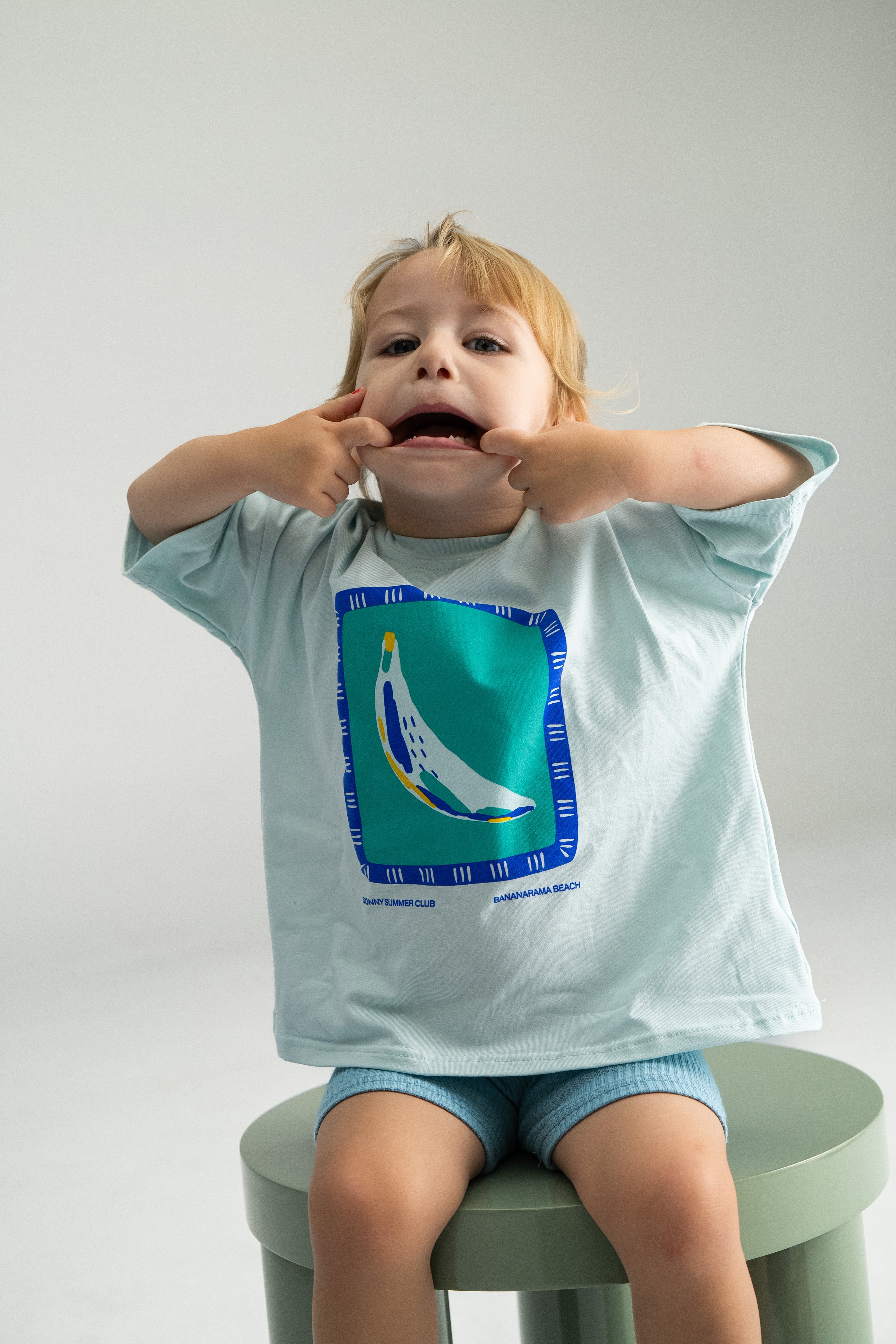 A child with light brown hair is sitting on a green stool, wearing the Bananarama Tee Baby Blue by SONNY LABEL. They are playfully making funny faces with their fingers in a bright, minimalist setting reminiscent of Bananarama Beach.