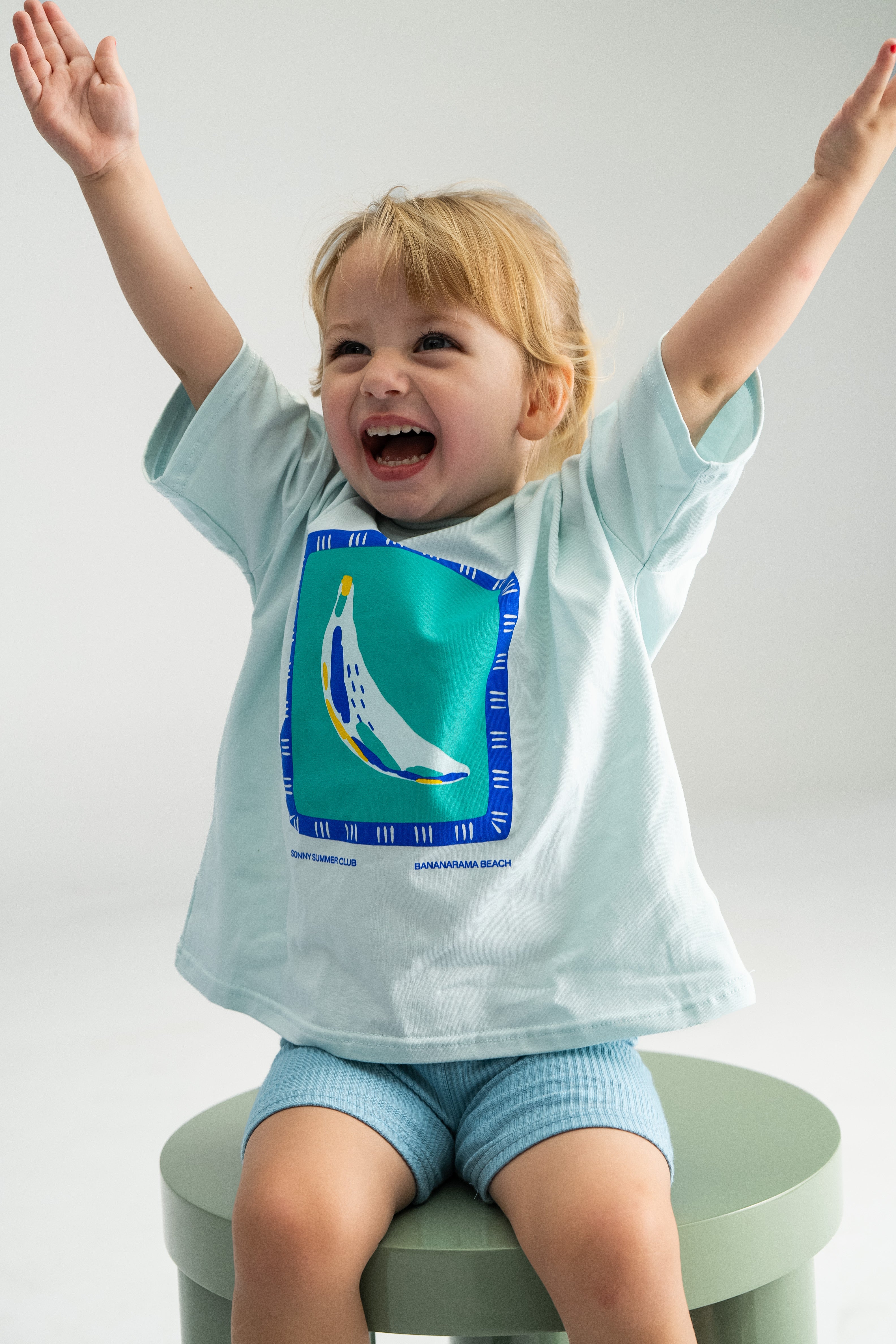 A joyful child with blonde hair wearing the Bananarama Tee Baby Blue by SONNY LABEL, featuring a colorful design, is sitting with arms raised on a green stool. The child is smiling widely against a plain white background.