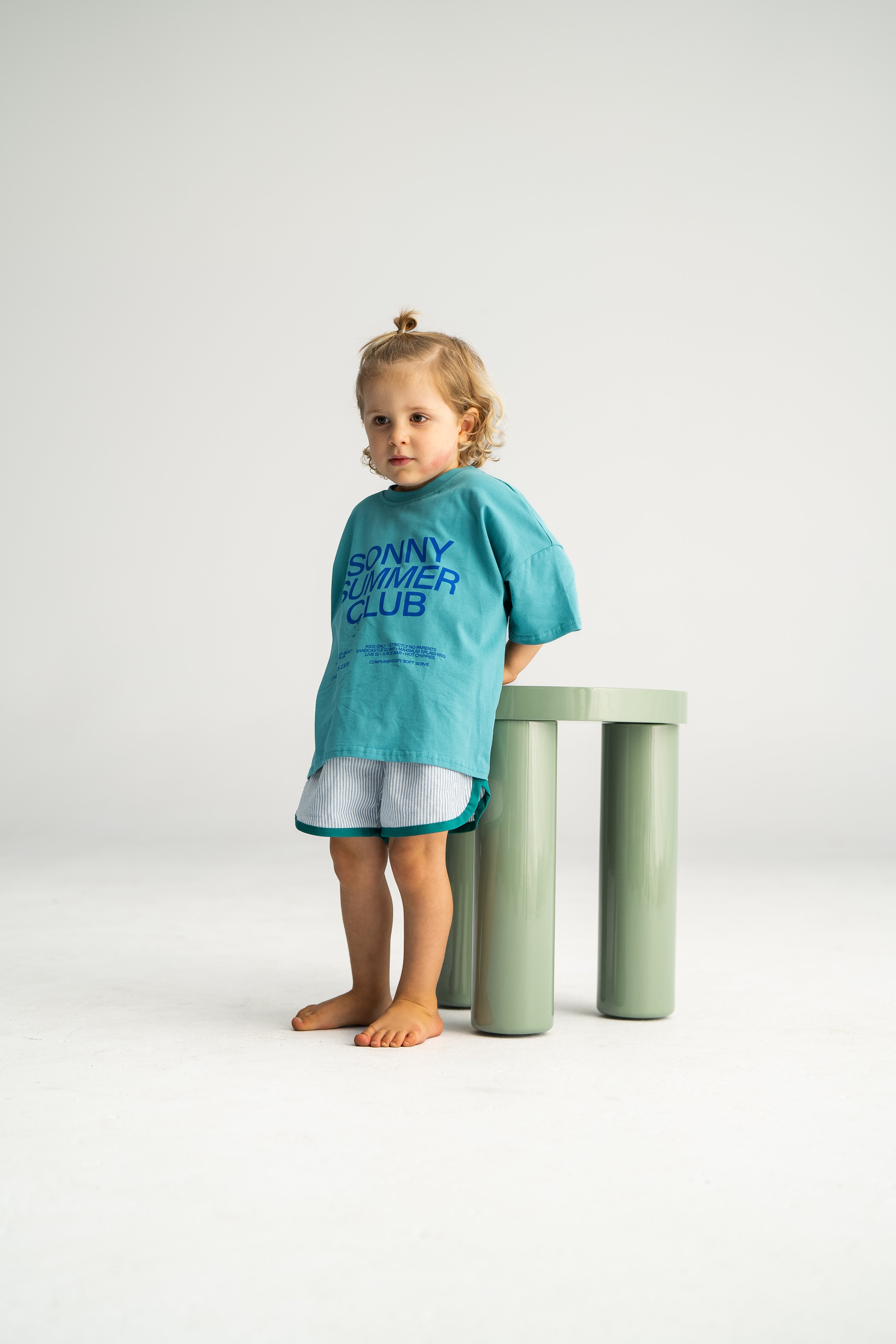 A barefoot child with a small topknot stands next to a light green cylindrical stool, wearing an oversized teal shirt from SONNY LABEL's "SONNY SUMMER CLUB" collection. They pair it with Stripe Shorties in blue, size 6, perfect for effortless summer comfort. The backdrop is plain white.