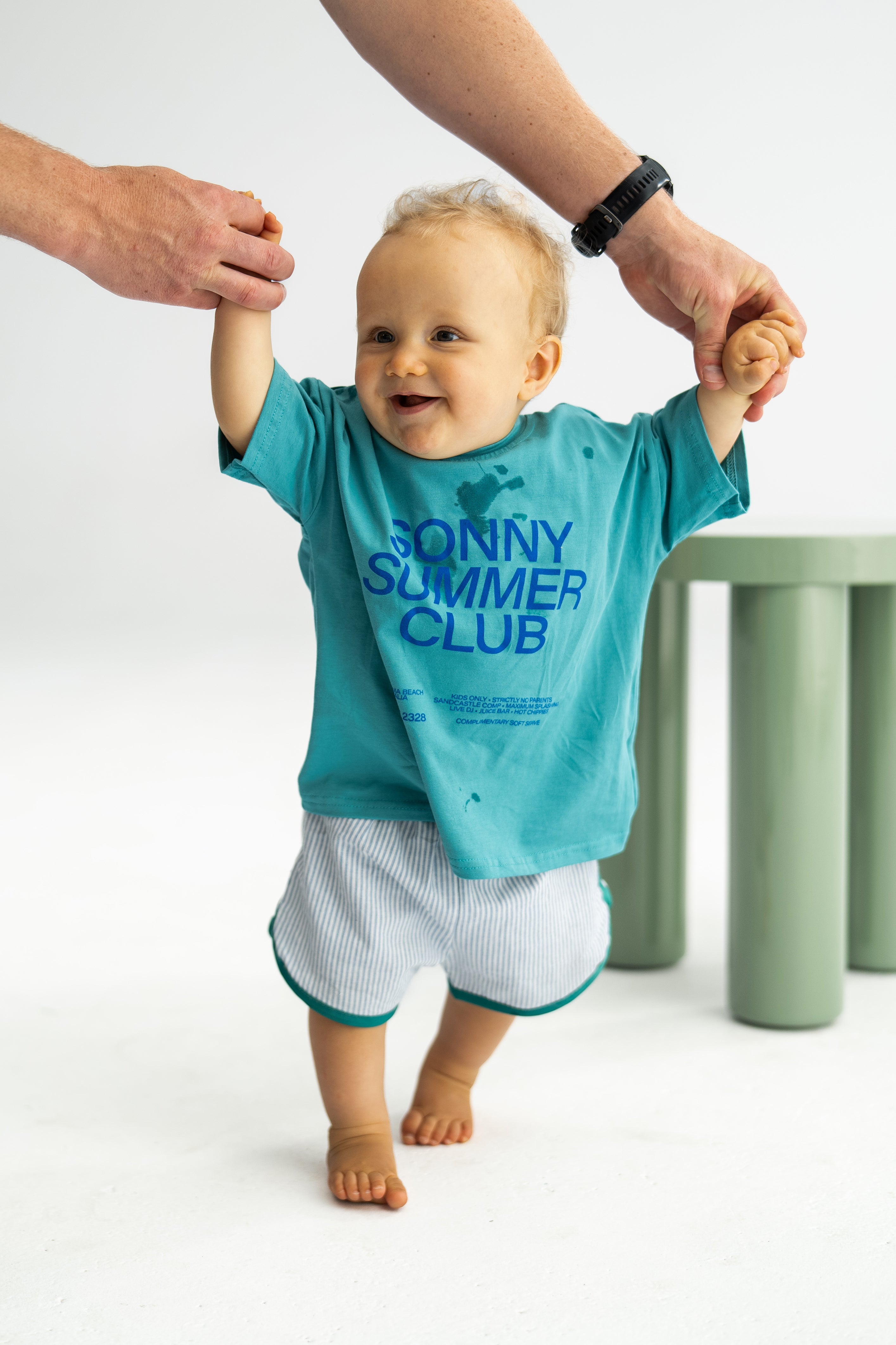 A smiling baby wears a turquoise "Sonny Summer Club" shirt and "Stripe Shorties Blue" by SONNY LABEL, supported by an adult's hands while taking steps. The baby enjoys summer comfort on a white floor with a light green table in the background.