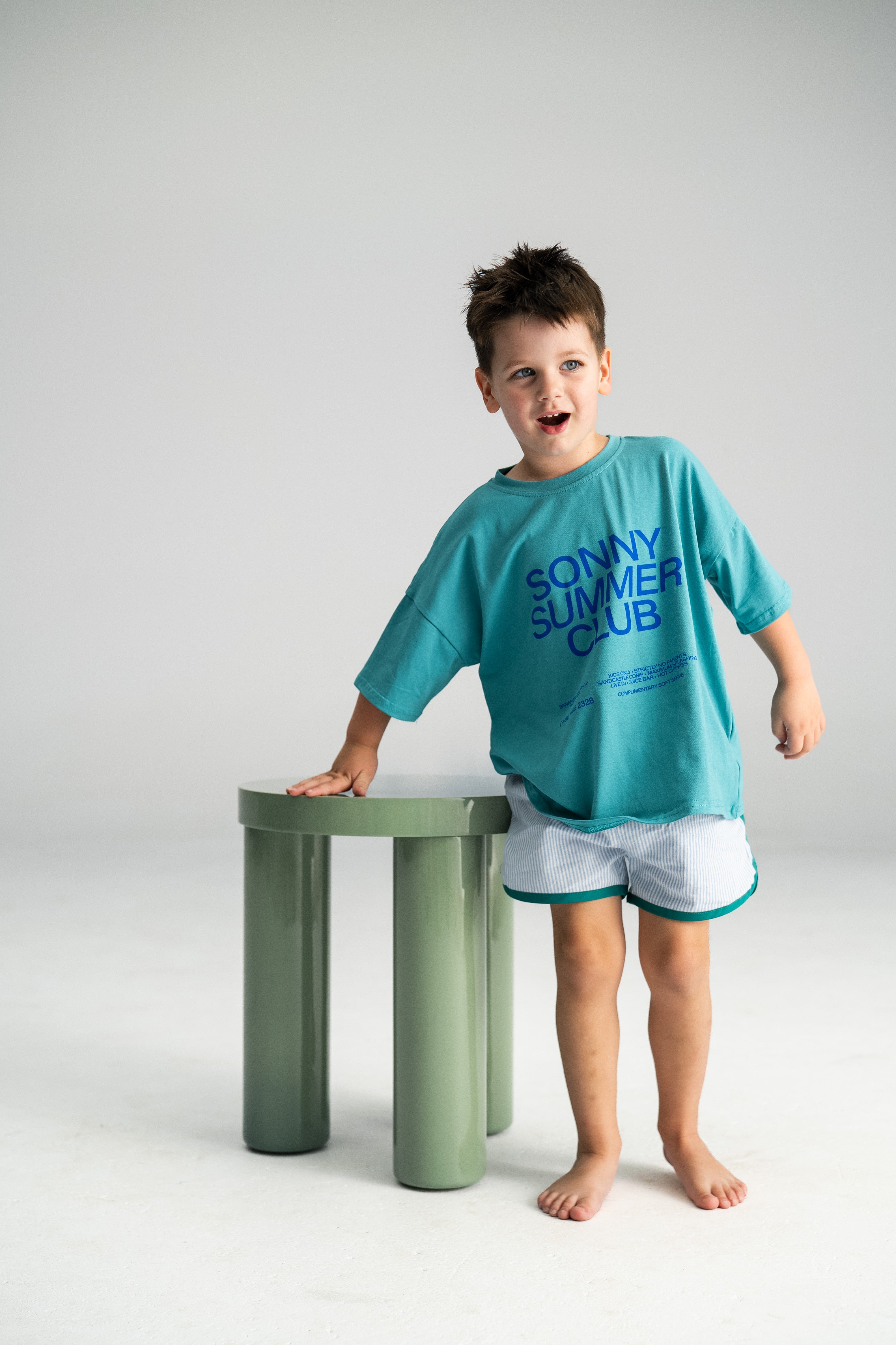 A young boy stands next to a small green table, embodying summer comfort in his SONNY LABEL 100% cotton blue t-shirt with "Sonny Summer Club" printed in purple and Stripe Shorties Blue - Size 6. With a playful expression, he's barefoot on a light background.