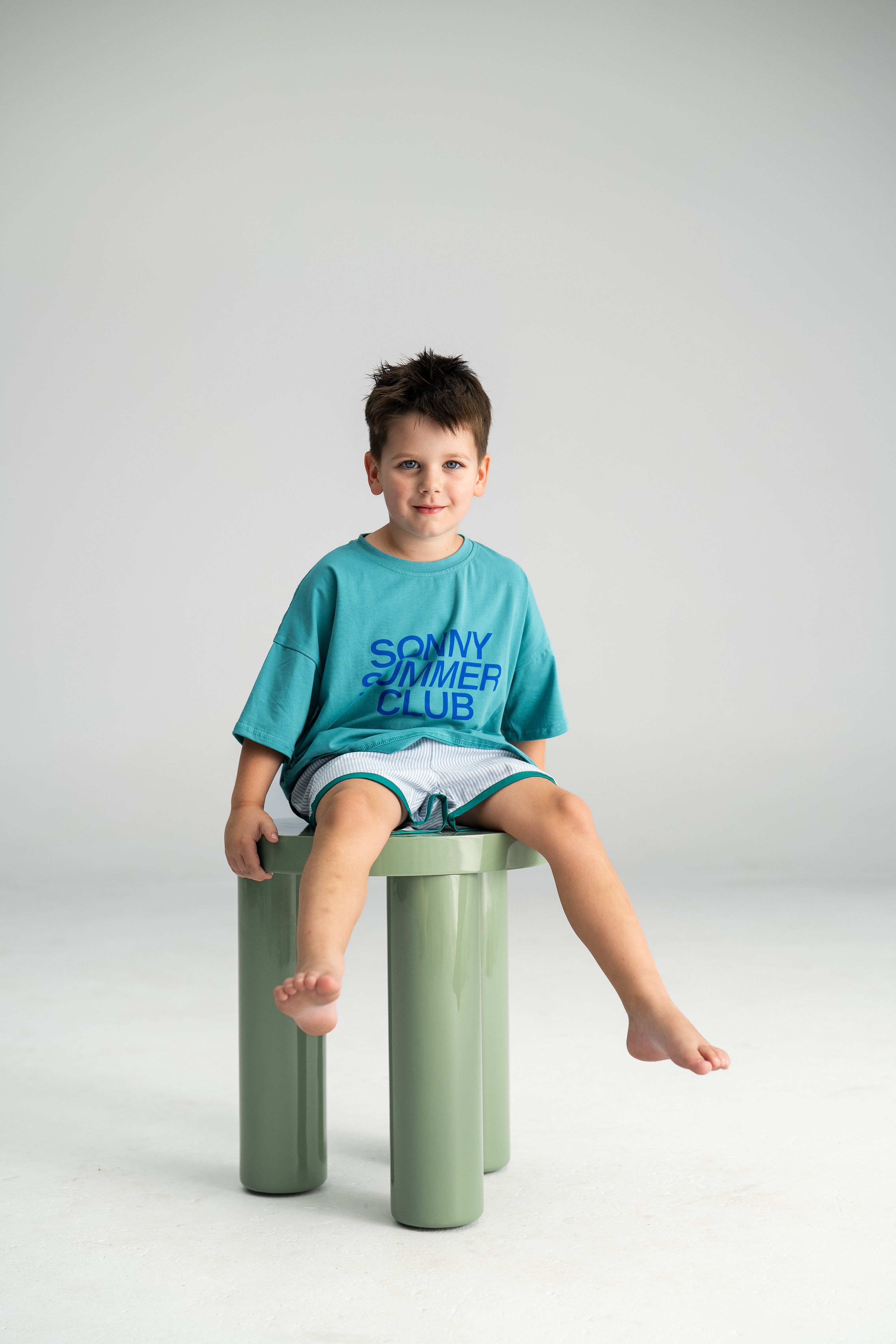 A young boy with short brown hair sits contentedly on a light green cylindrical stool. He's wearing a teal T-shirt with "SONNY SUMMER CLUB" printed on it and lightweight gray shorts, specifically the Stripe Shorties Blue by SONNY LABEL, for ultimate summer comfort. The background is a plain, light gray.