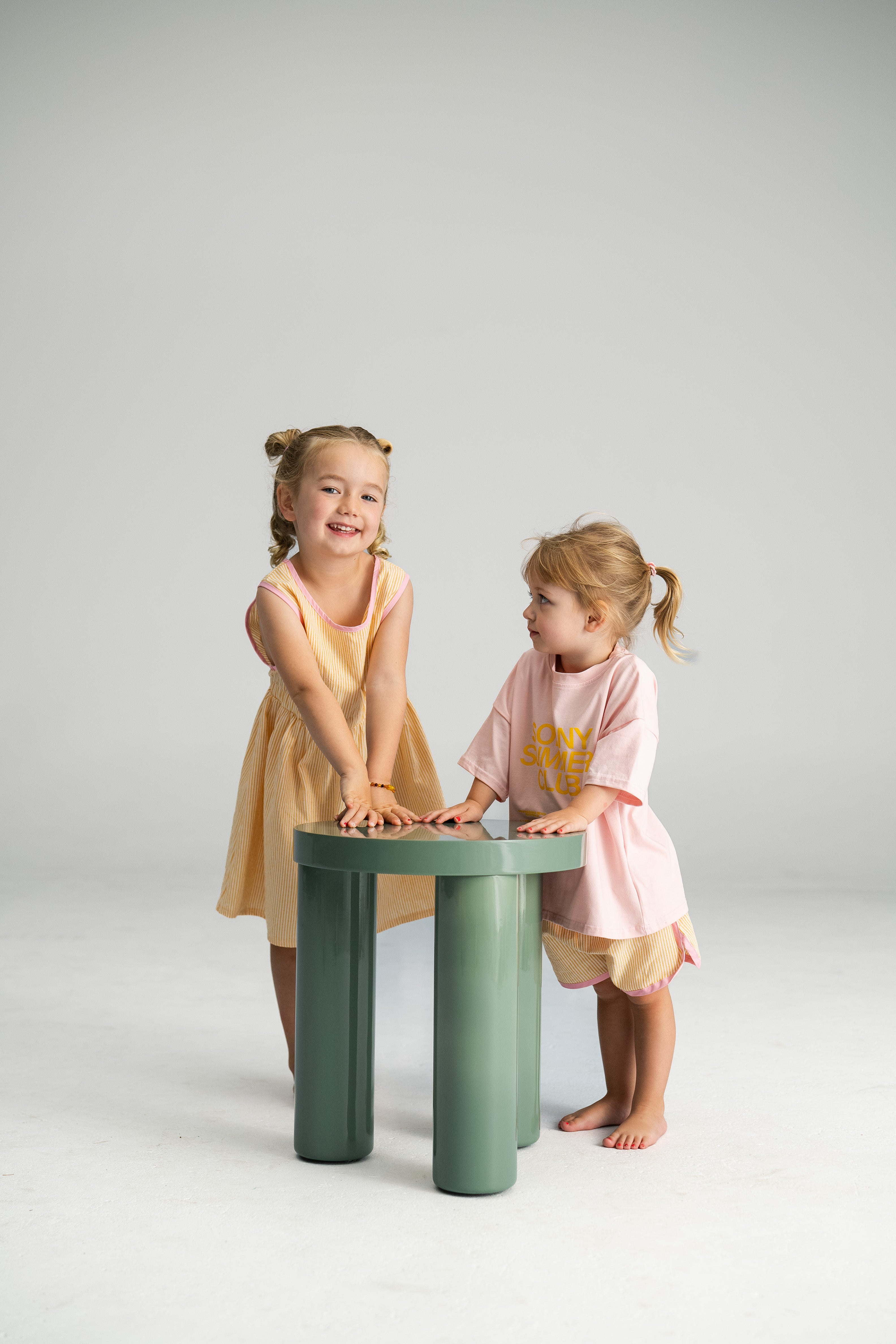 Two young children stand by a small green table in a bright room. One child, wearing the Stripe Shorties Lemon from Sonny Label, smiles at the camera. The other, dressed in a pink shirt with contrast binding, looks towards the first child. Both have their hair styled in small ponytails.