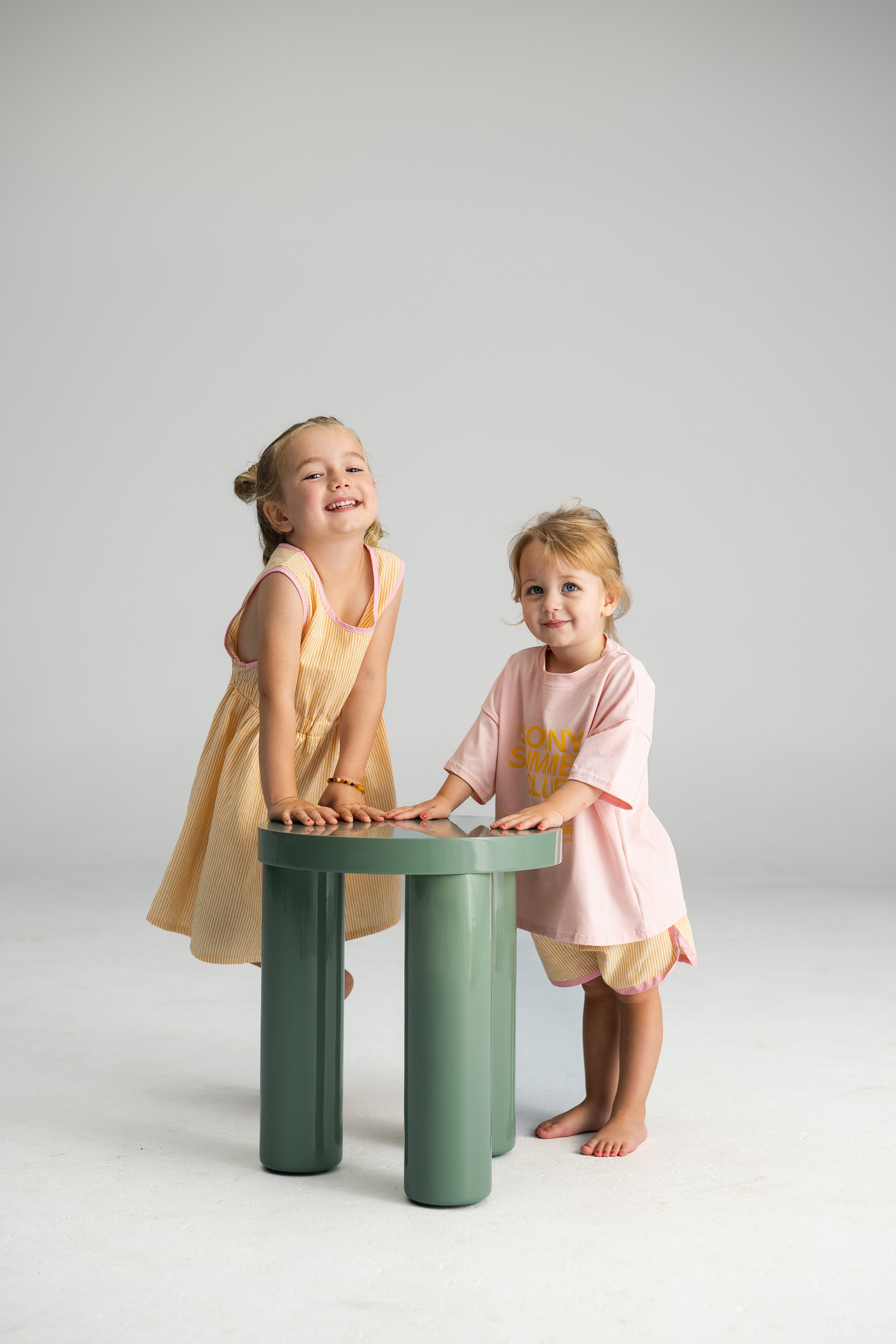 Two young children are smiling and leaning on a small green table. The child on the left is wearing a yellow dress, while the child on the right sports SONNY LABEL's Stripe Shorties Lemon—a pink shirt with yellow shorts featuring pink contrast binding. The background is plain and light-colored, perfect for showcasing summer comfort.