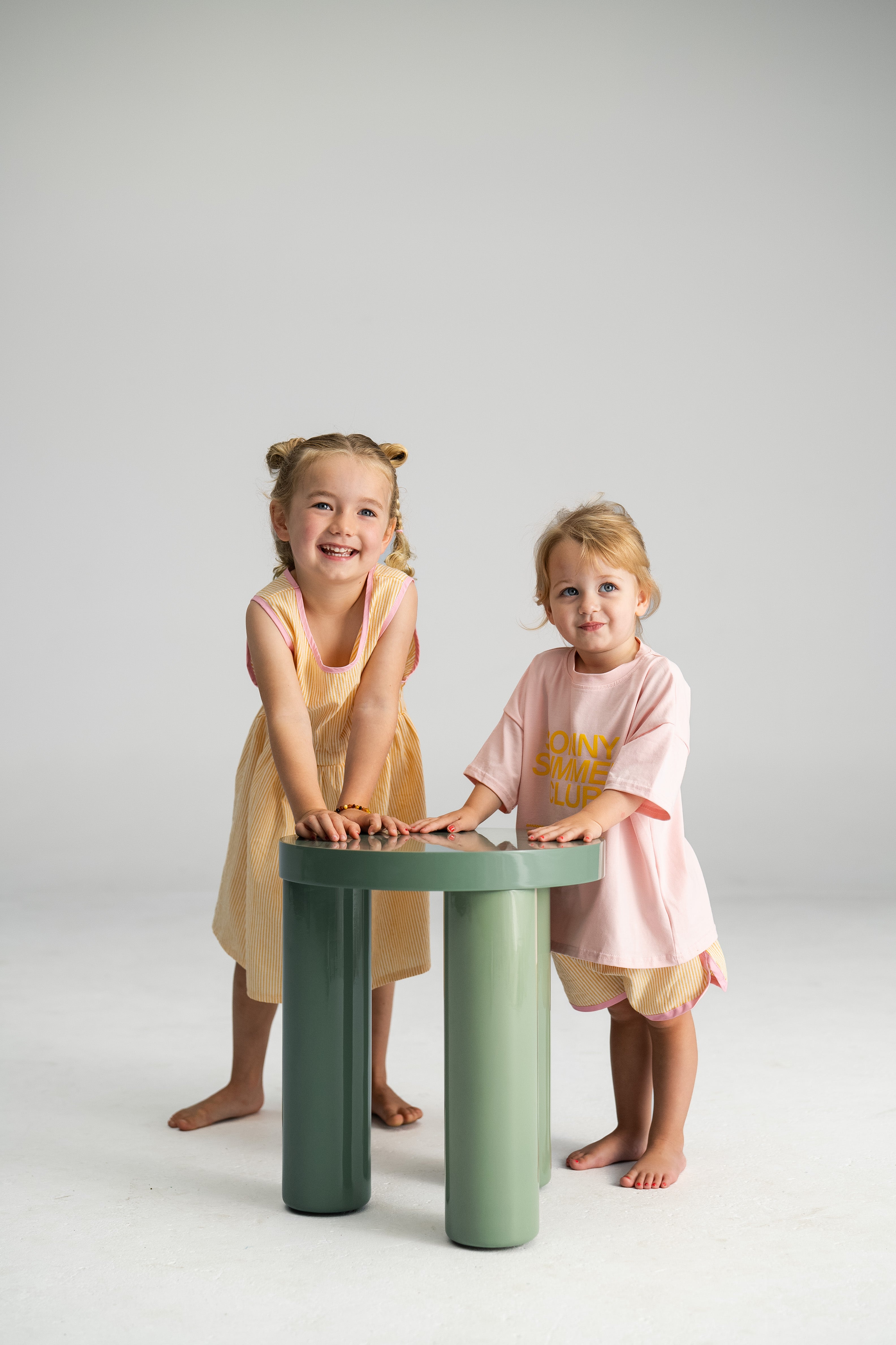 Two young children stand next to a green round table. The girl is dressed in the Stripe Pinafore Lemon from SONNY LABEL, made from 100% cotton, while the boy wears a pink shirt and shorts. Both are smiling and leaning on the table against a plain background.