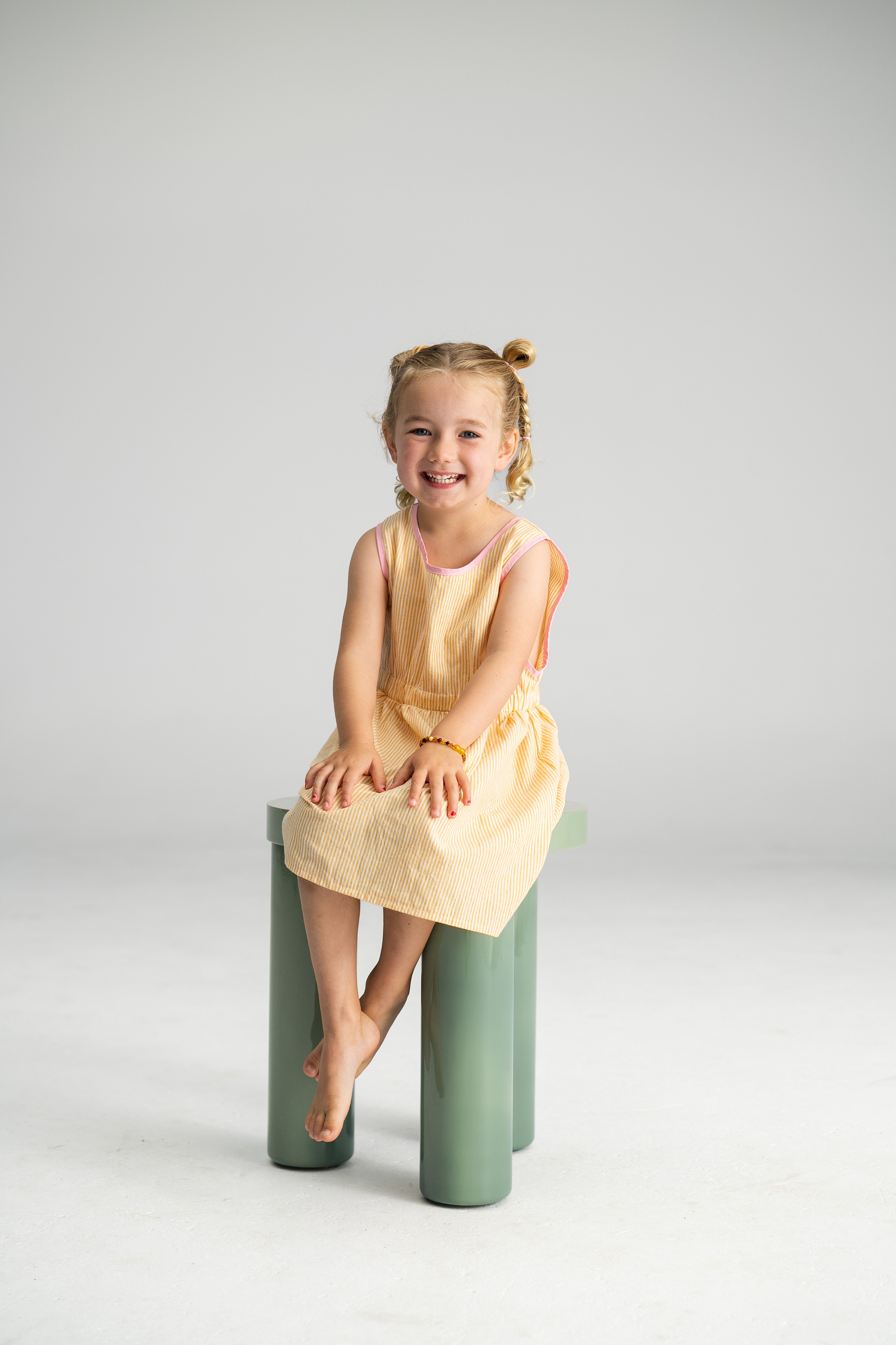 A young girl with braided hair sits smiling on a green stool, her SONNY LABEL Stripe Pinafore in lemon adding a touch of charm. Barefoot and carefree, the plain, light-colored background perfectly highlights her cheerful expression in this 100% cotton delight.