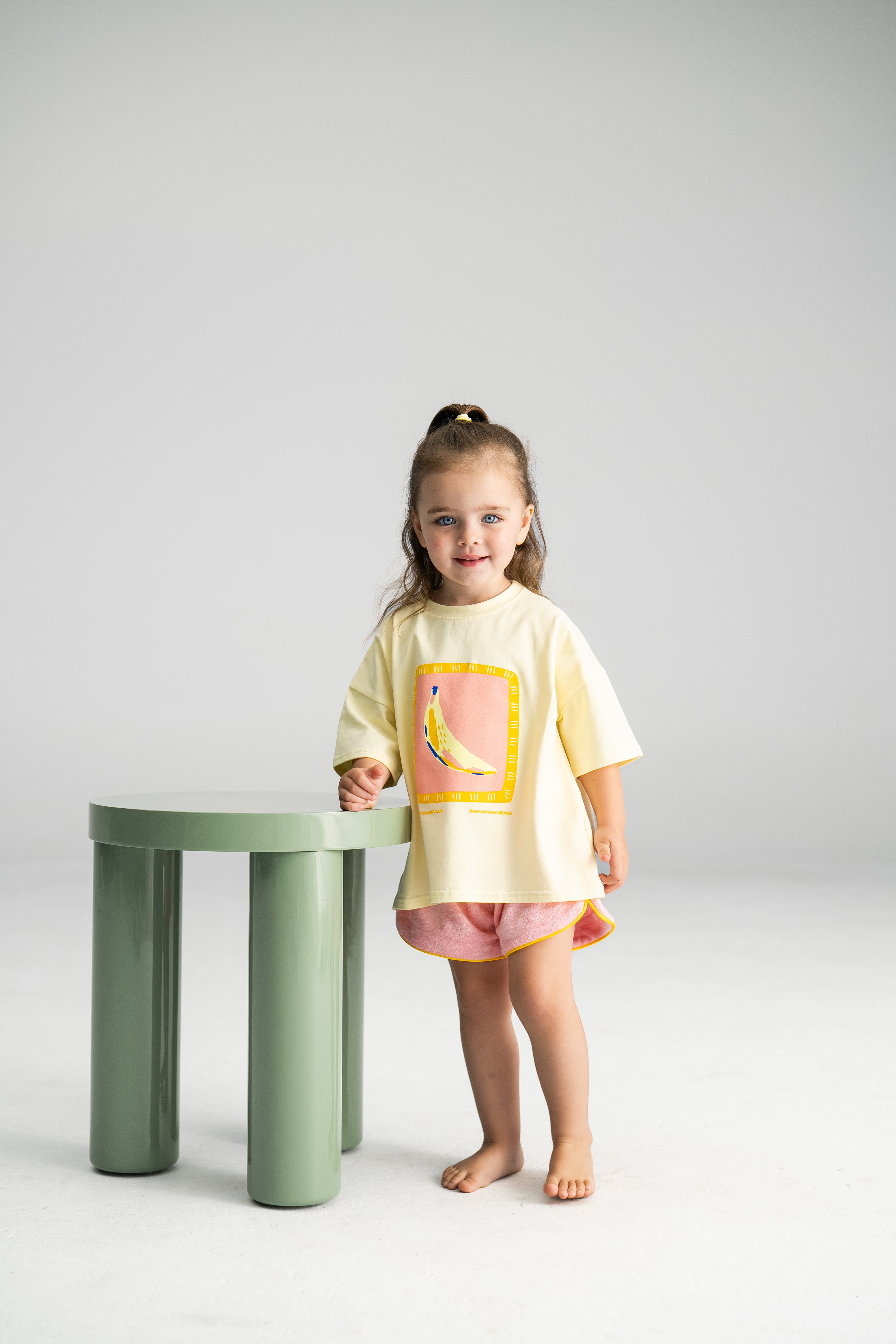 A young child with long brown hair in a ponytail stands barefoot beside a small green table. They're wearing a yellow top featuring a colorful banana graphic and SONNY LABEL's Terry Shorties in pink. The light and minimalistic background enhances the cheerful vibe of the outfit.