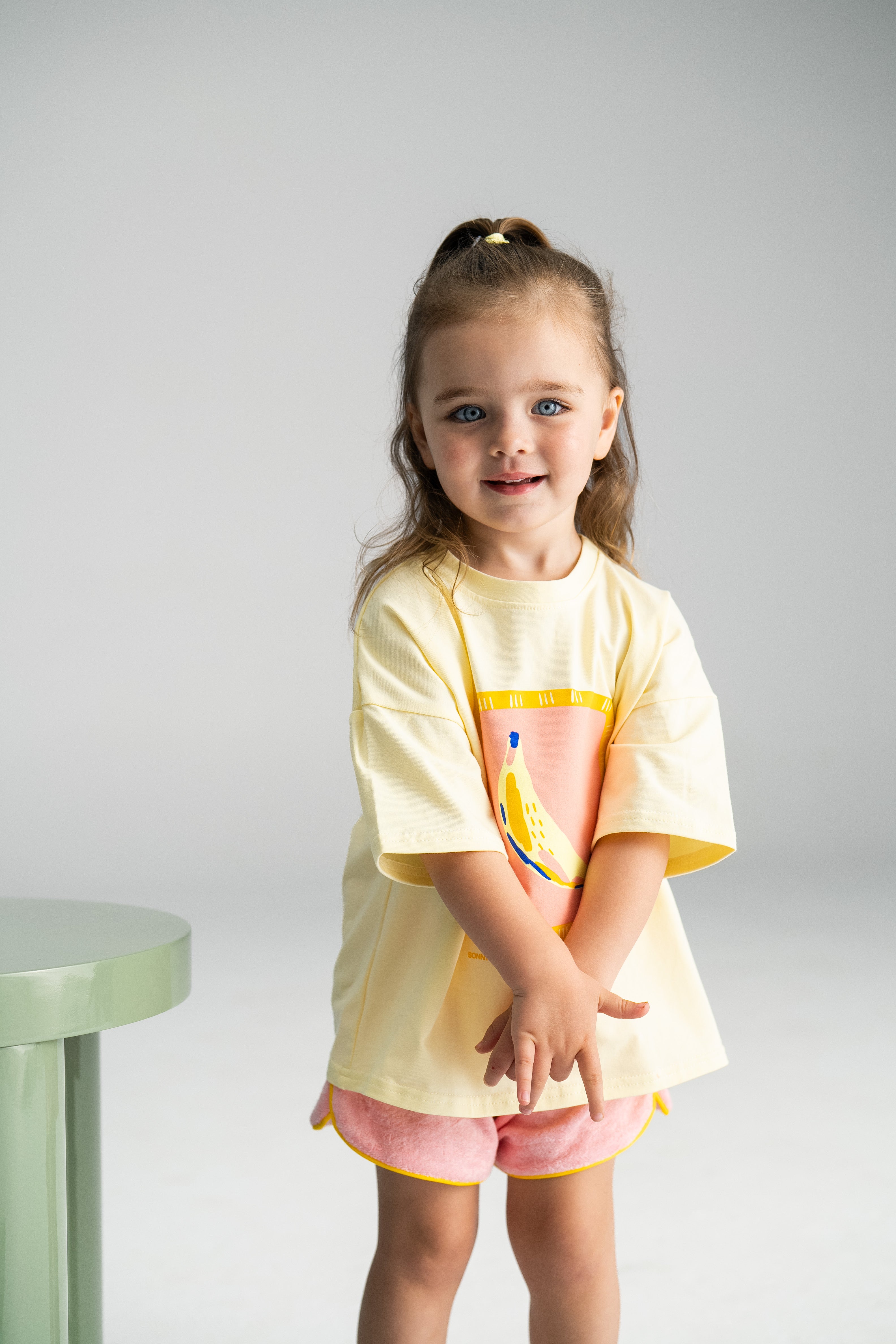 A young girl with long brown hair in a ponytail is standing and smiling. She is wearing a yellow t-shirt with a colorful bird design and the pink Terry Shorties from SONNY LABEL. Her hands are clasped together in front of her, and a small green table is to her left.