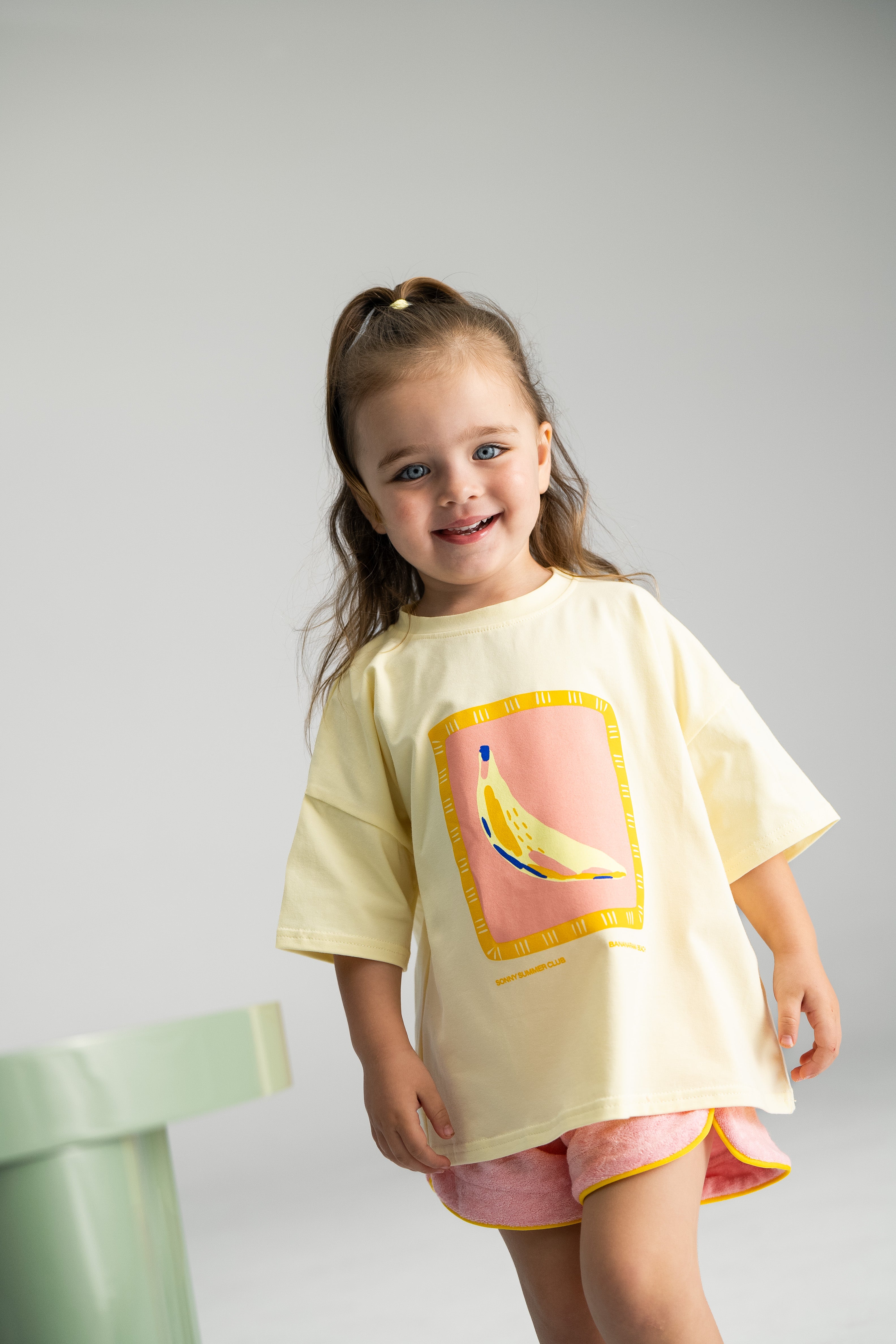 A young child with long hair grins, dressed in a yellow, loose-fit T-shirt adorned with a banana graphic. The outfit is complemented by the Terry Shorties Pink from SONNY LABEL, while the child stands in a brightly lit room with a light green object partially visible in the foreground.